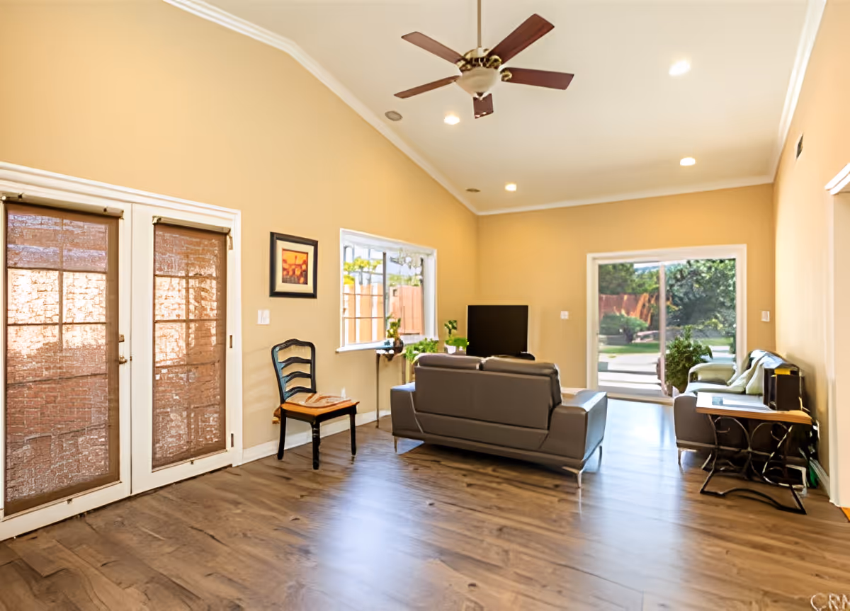 A spacious living room with beige walls and wooden flooring. The room features a ceiling fan with lights, a gray sofa facing a flat-screen TV, a wooden chair with a cushion, a small table with plants near a window, and a sliding glass door leading to an outdoor area with greenery. There is also a small desk with a chair and some items on it along the right wall.