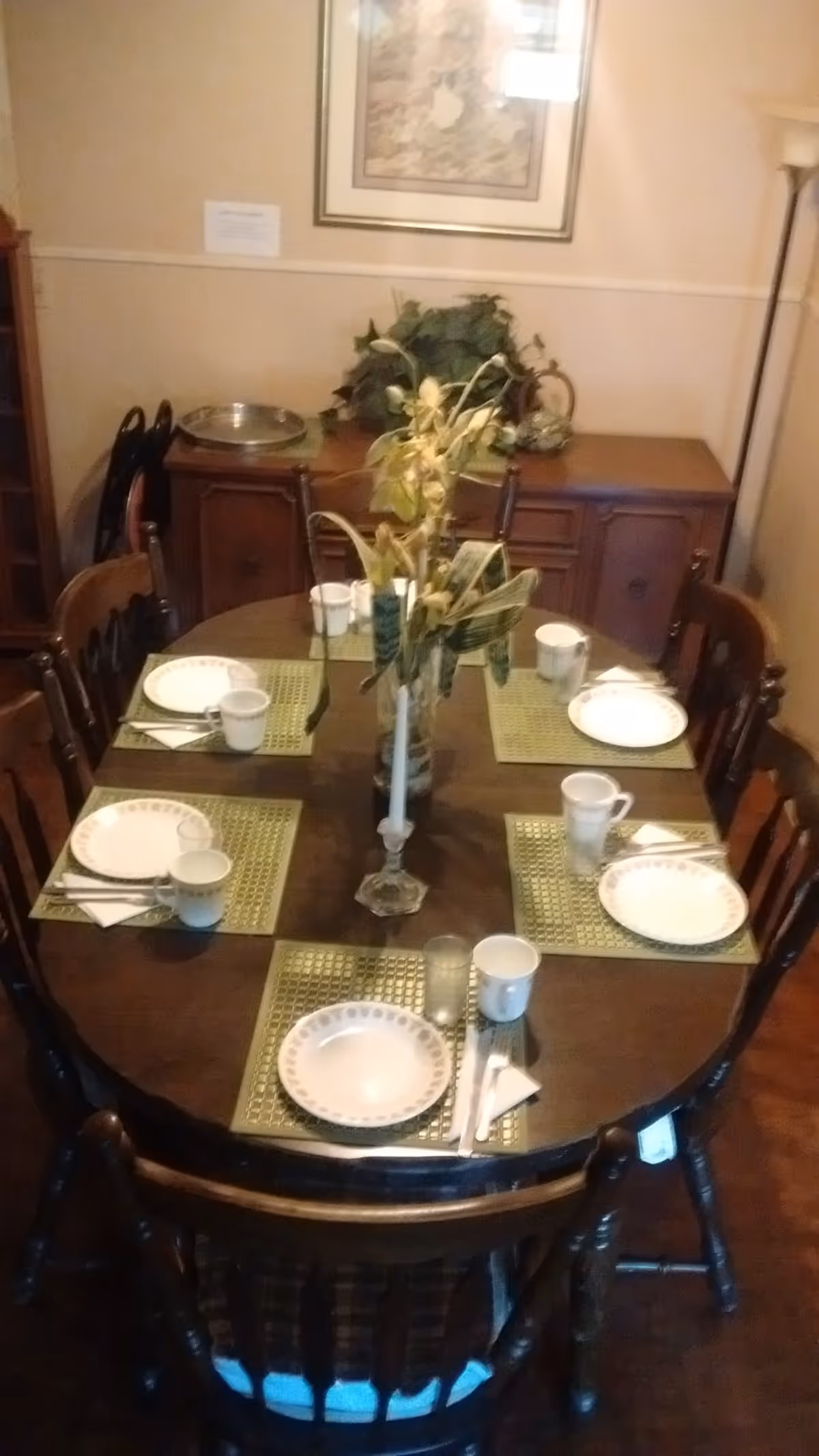 A dining room with a wooden oval table set for six people. Each place setting includes a white plate with a decorative border, a cup, a glass, and silverware on a green placemat. A vase with dried flowers and a white candle in a holder are placed in the center of the table. In the background, there is a wooden sideboard with a silver tray and a leafy green plant. A framed picture hangs on the wall above the sideboard, and a floor lamp is visible on the right side.