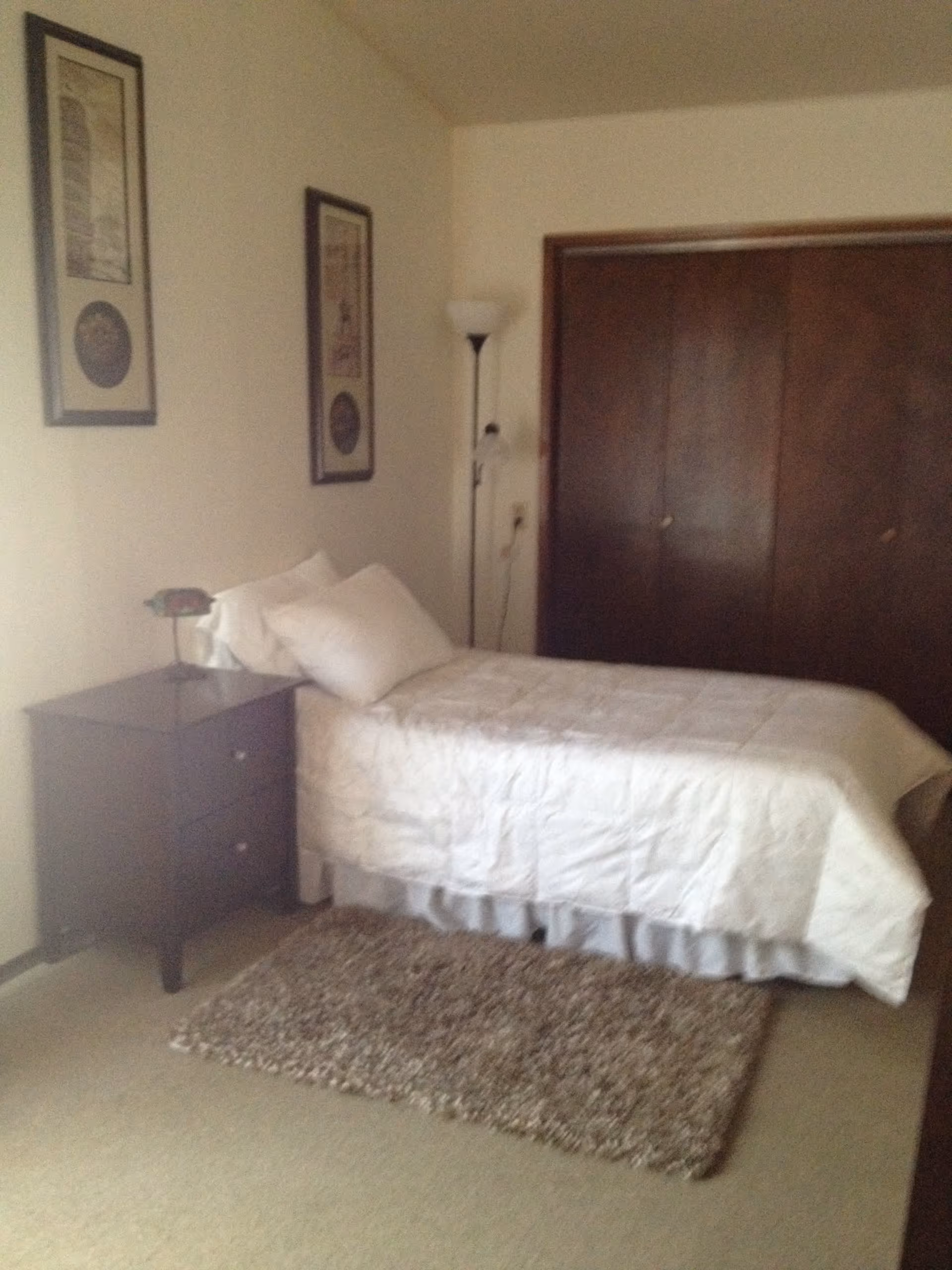 A simple bedroom with a single bed covered in a white quilt and pillows. Next to the bed is a dark wooden nightstand with a small lamp. There is a tall floor lamp in the corner near a large wooden closet with sliding doors. Two framed pictures hang on the wall above the bed. A textured rug is placed on the floor beside the bed.