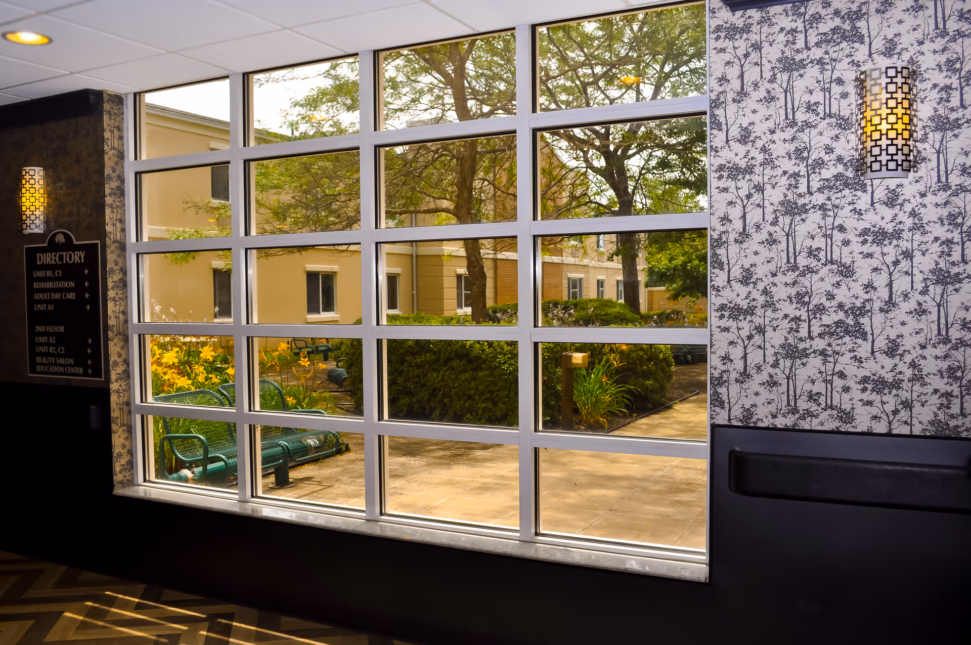 View through a large multi-pane window showing an outdoor garden area with green benches, yellow flowers, bushes, and trees. Inside, there is patterned wallpaper, a wall sconce light, and a directory sign on the left side.