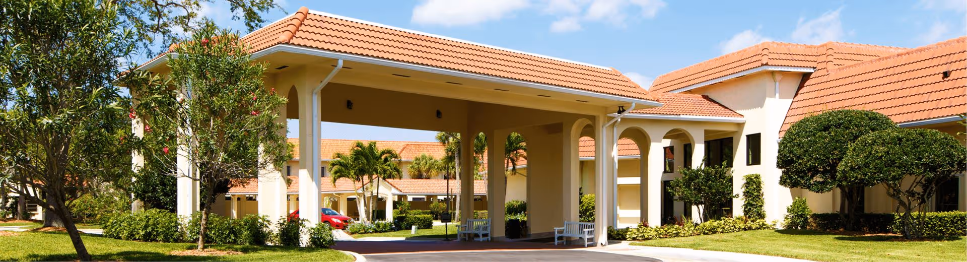 Covered porte-cochère entrance with tiled roofs and landscaped grounds in front of a senior living facility.