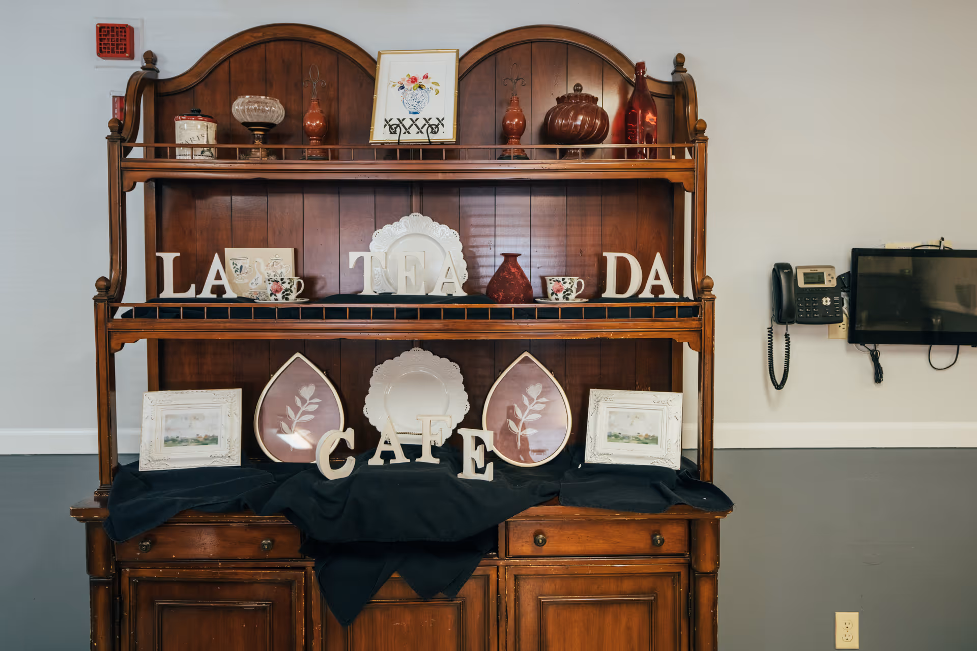 A wooden hutch displaying decorative plates, teacups, framed pictures, and letters spelling out 'LA TEA DA CAFE'. The hutch is set against a plain wall with a mounted telephone and a small television screen on the right side.
