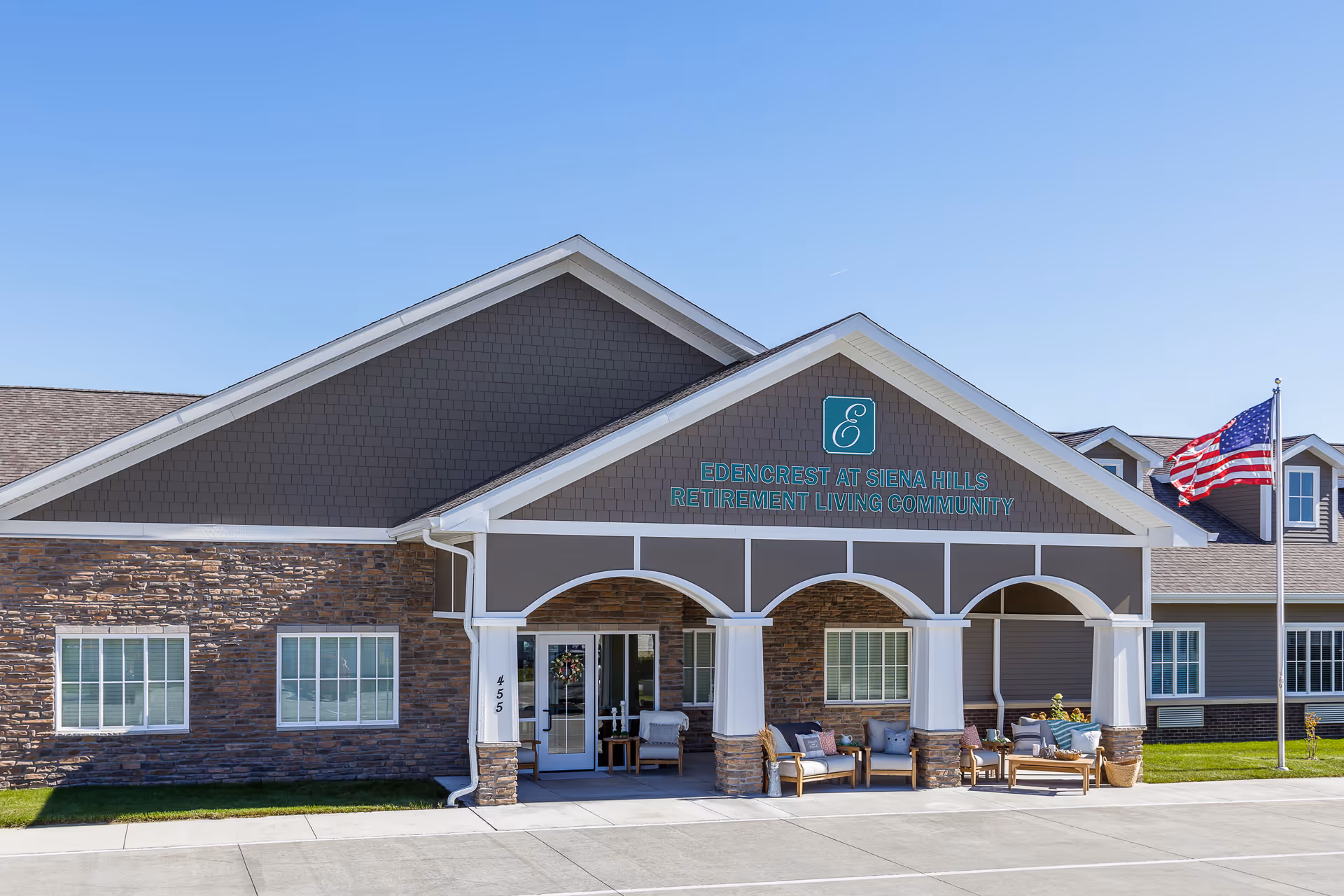 Front exterior view of Edencrest at Siena Hills retirement living community building with a covered entrance, outdoor seating, and an American flag on a flagpole under a clear blue sky.