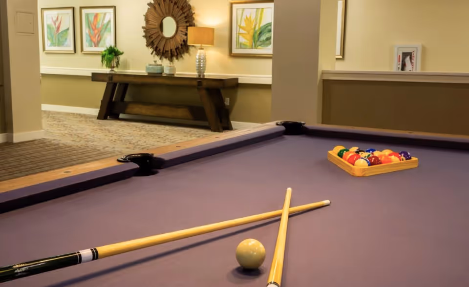 A pool table with a purple felt surface, two crossed pool cues, a white cue ball, and a rack of colorful billiard balls. In the background, there is a wooden console table with a lamp, decorative items, a round mirror, and framed botanical artwork on the wall.