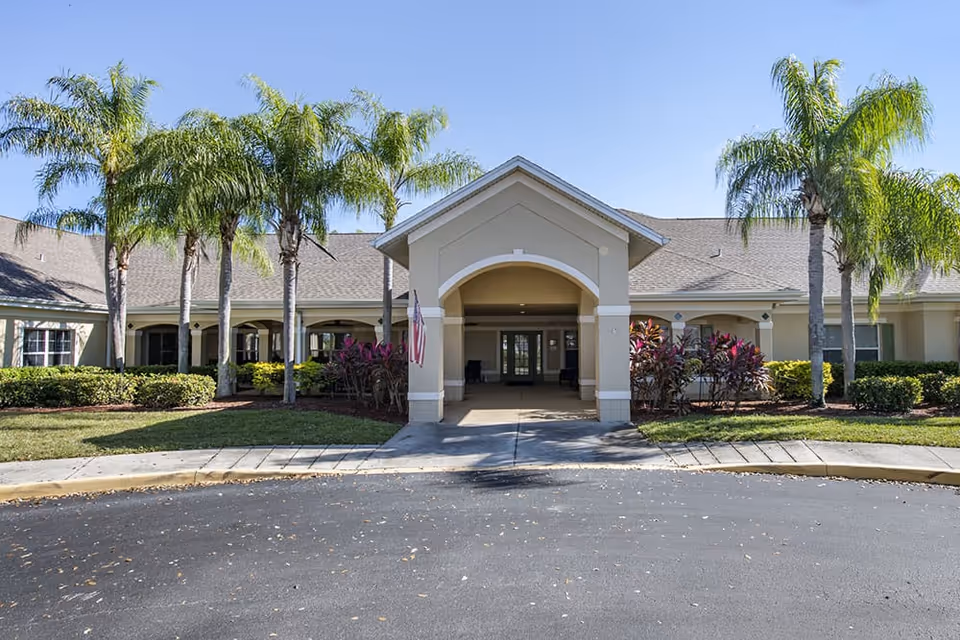 Front exterior view of a single-story senior living facility building with a covered entrance, palm trees, and well-maintained landscaping under a clear blue sky.