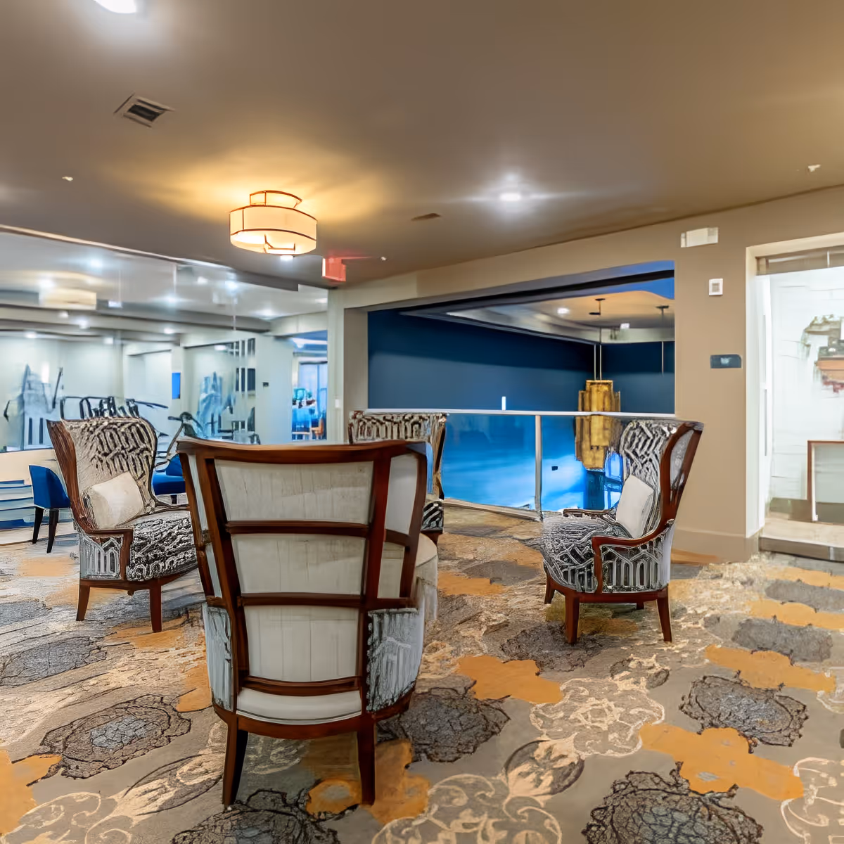 A comfortable communal lounge with upholstered armchairs arranged on a patterned carpet overlooking an indoor pool area.