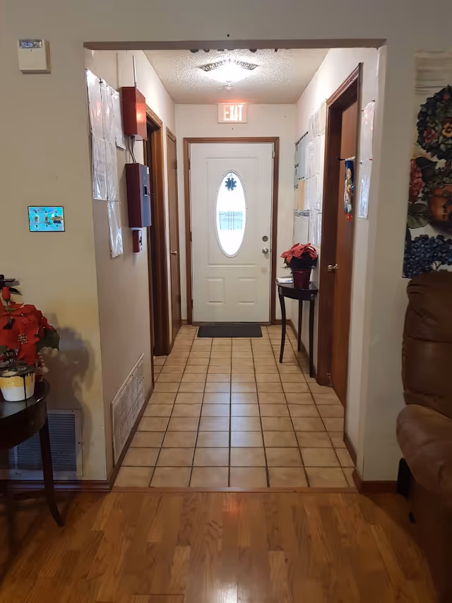 Interior hallway with a tiled floor leading to a white front door beneath an EXIT sign, flanked by doors and small tables with poinsettias.