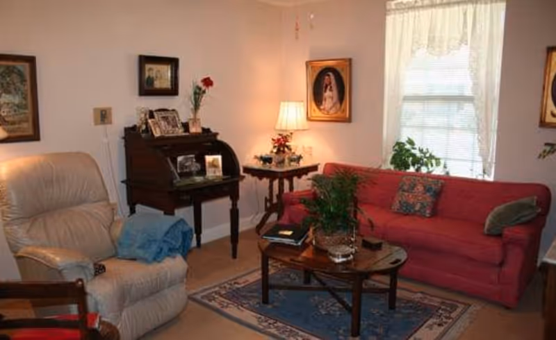 A cozy living room with a red sofa adorned with two pillows, a beige recliner chair with a blue blanket draped over it, a wooden coffee table with a plant and books, a small side table with a lamp and decorative items, and framed pictures hanging on the walls. A window with sheer curtains allows natural light into the room.