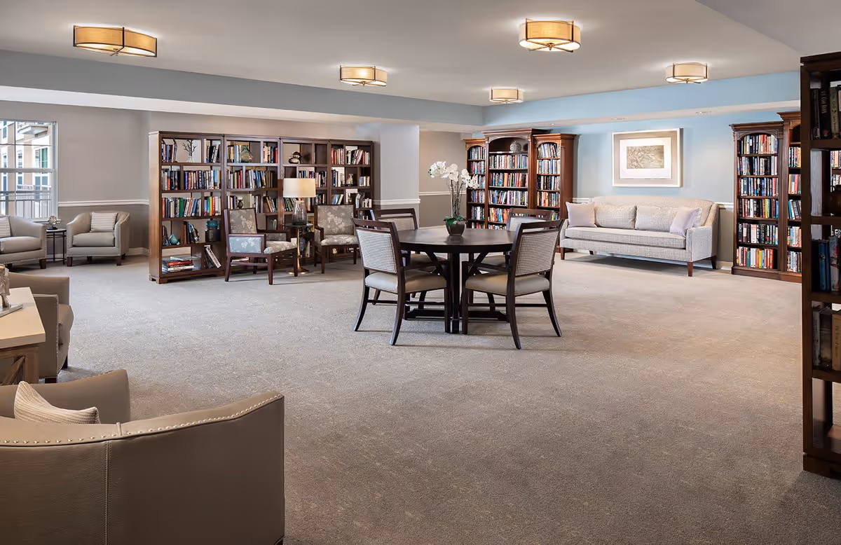 A spacious senior living common area with beige carpeting, multiple wooden bookshelves filled with books, a round table with four chairs in the center, several armchairs and a sofa along the walls, soft overhead lighting, and a framed picture on the wall.