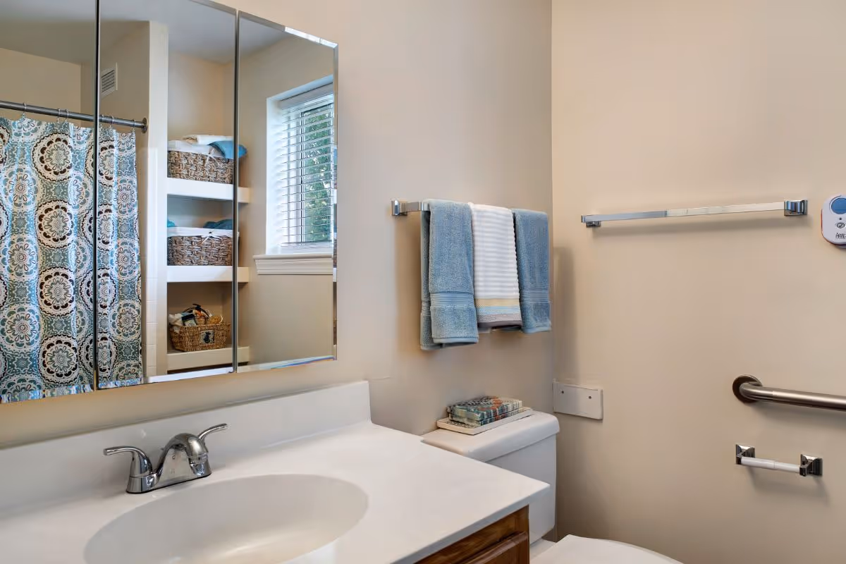 A clean bathroom with a white sink and countertop, a silver faucet, a mirror cabinet above the sink, a window with blinds, shelves with wicker baskets and towels, a toilet with folded towels on top, and a towel rack with blue and white towels hanging. There is also a patterned shower curtain visible in the mirror reflection.