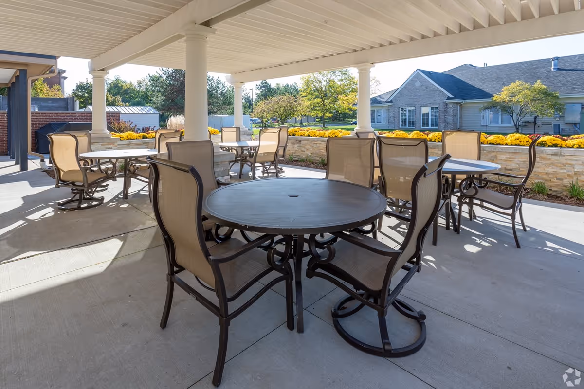 Outdoor covered patio area with multiple round tables and beige mesh chairs arranged around them. The patio has white columns supporting the roof and overlooks a garden with yellow flowers and trees. Residential buildings are visible in the background under a clear sky.