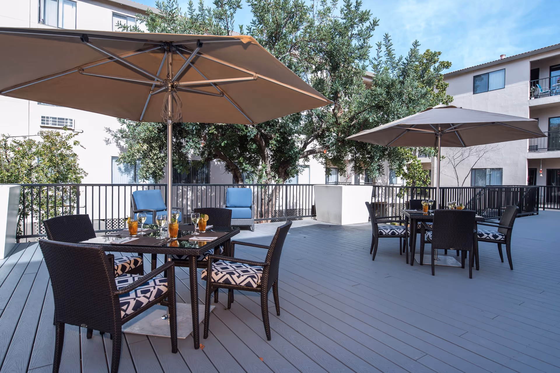 Outdoor patio area with two tables, each surrounded by four chairs with patterned cushions. Each table has a large beige umbrella providing shade. There are drinks on the tables and two blue cushioned chairs near the railing. Trees and a building with balconies are visible in the background under a clear blue sky.