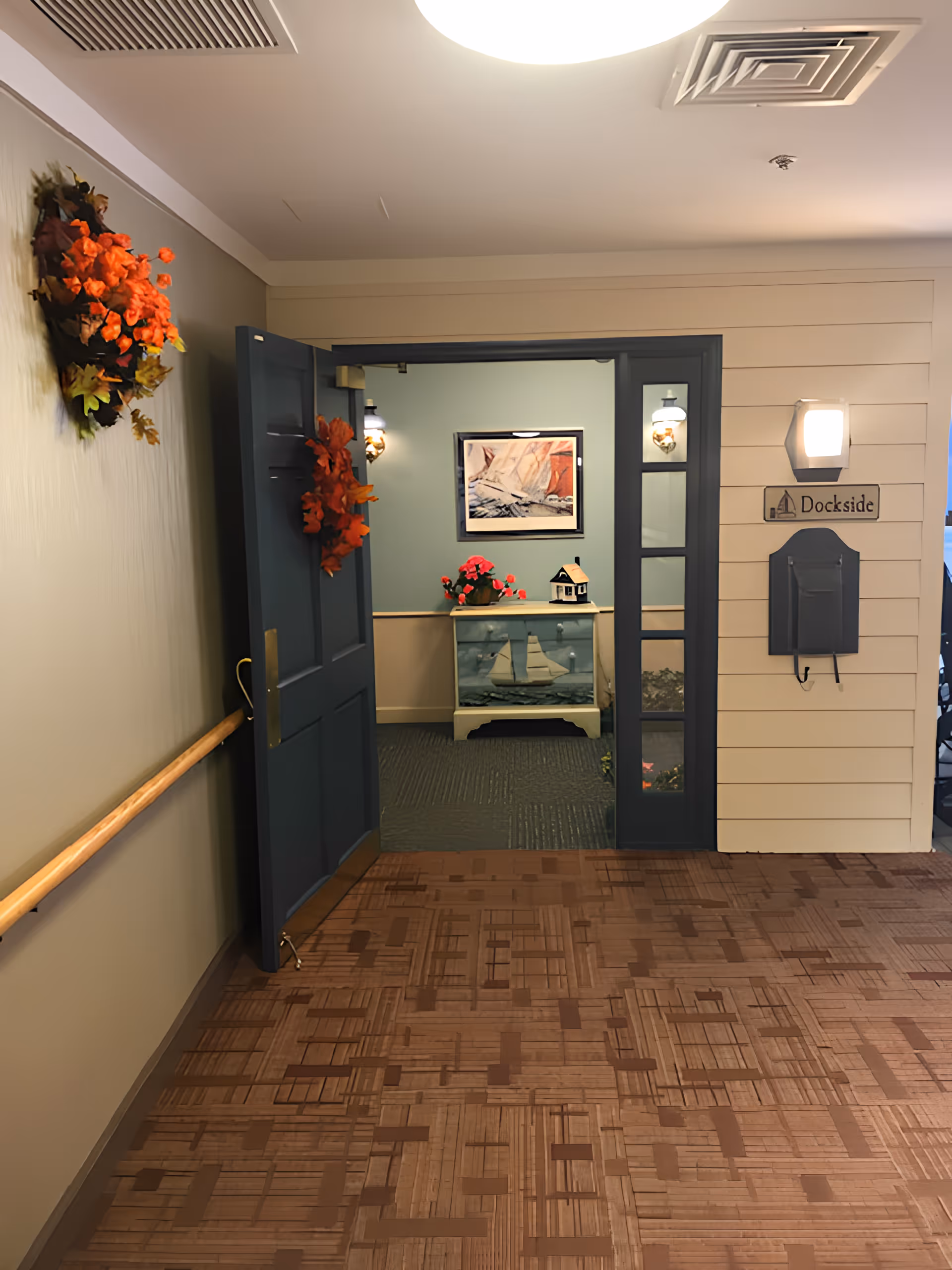Interior hallway of a senior living facility with a blue door open to a room labeled 'Dockside'. The hallway has a wooden handrail on the left wall and a fall-themed wreath with orange leaves. Inside the room, there is a small cabinet with a sailboat decoration, a flower pot with red flowers, and a framed picture on the wall. The floor has a patterned carpet and the walls are painted in neutral tones.