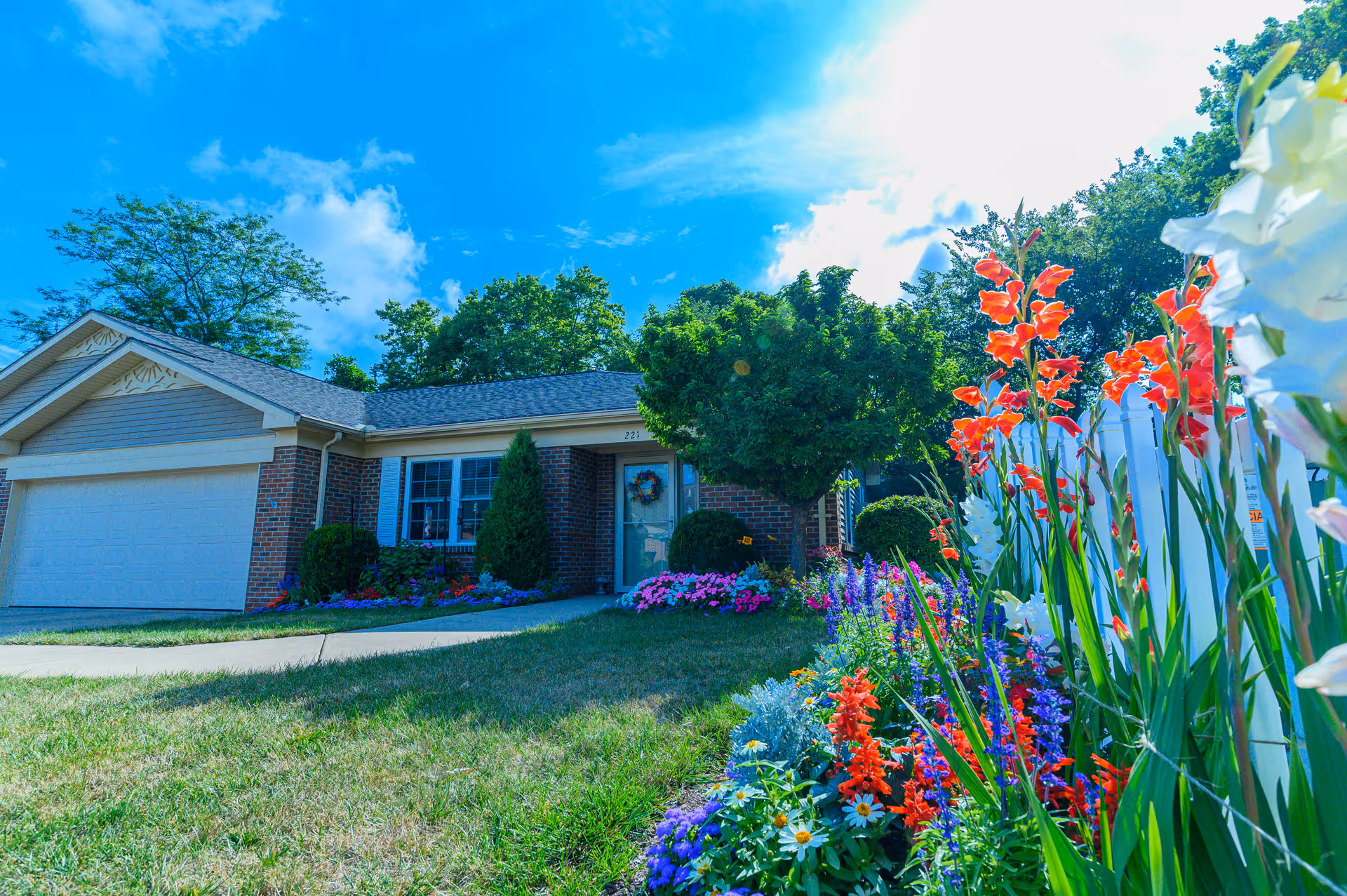 A single-story brick house with a two-car garage, surrounded by a well-maintained lawn and colorful flower beds along a white picket fence under a bright blue sky with some clouds.