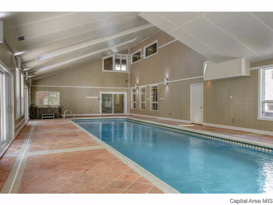 Indoor swimming pool in a high-ceiling room with tiled deck and large windows.