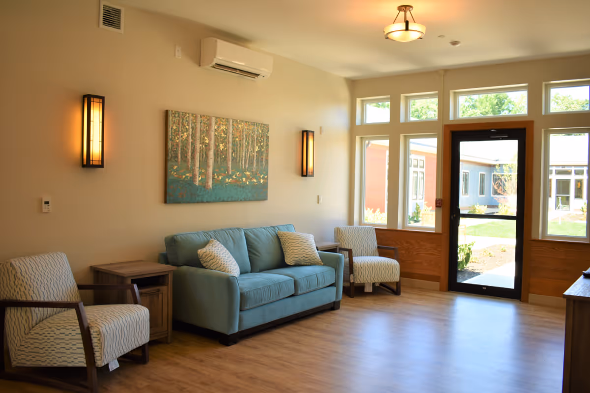 A bright and cozy living room with a light blue sofa, two patterned armchairs, a wooden side table, and a painting of a forest on the wall. The room has large windows and a glass door that leads outside, allowing natural light to fill the space. Warm wall sconces provide additional lighting.