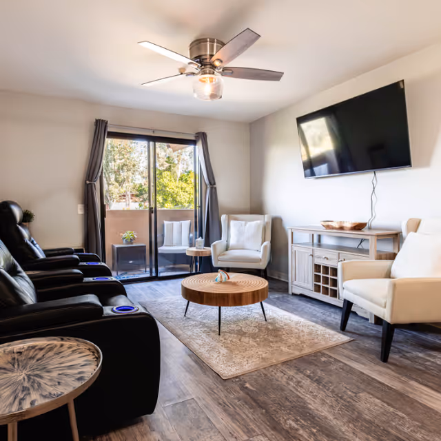 A bright living room with a ceiling fan, three black leather recliners with cup holders, two white armchairs, a round wooden coffee table on a patterned rug, a wooden console with a mounted flat-screen TV above it, and sliding glass doors leading to a small outdoor patio with two chairs and a small table.