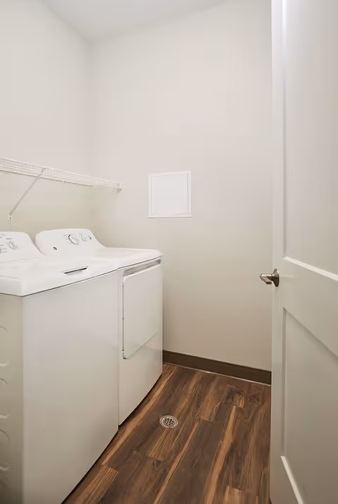 A small laundry room with a white washing machine and dryer side by side, a white wire shelf mounted on the wall above them, light beige walls, and wood-patterned flooring. The door to the room is partially open.