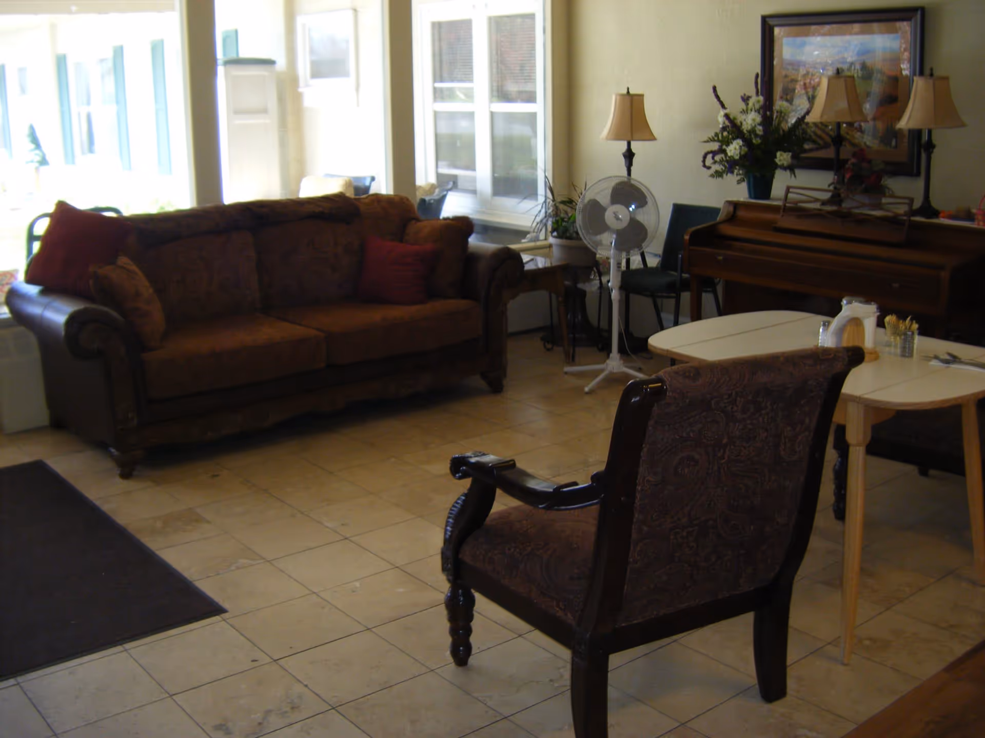 A common room featuring a sofa, upholstered armchair, small tables, a piano and a floor fan by large windows.