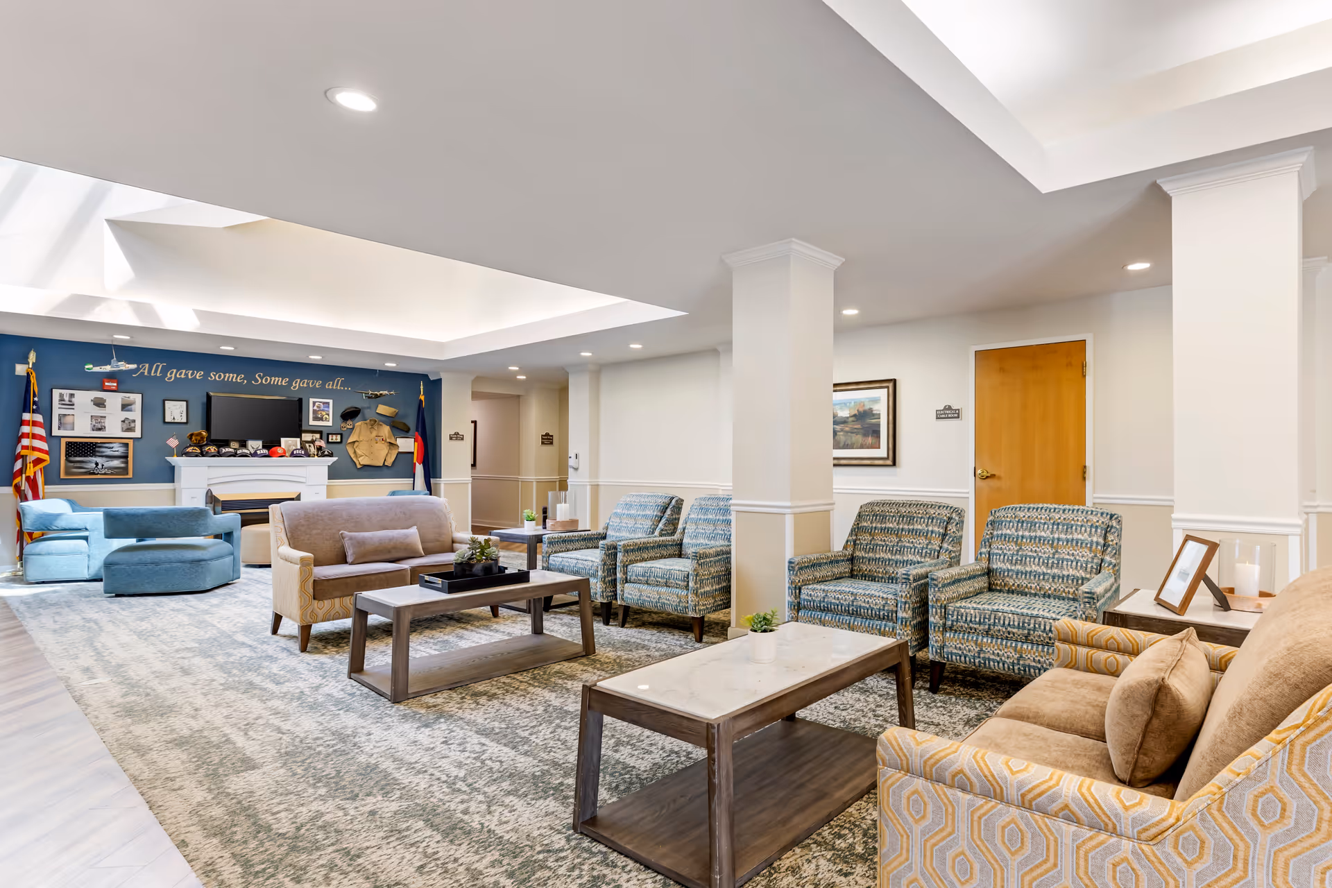 A spacious and well-lit living room area in a senior living facility with multiple upholstered chairs and sofas arranged around wooden coffee tables. The room features a blue accent wall with a display of military memorabilia, an American flag, and a Colorado state flag. The ceiling has recessed lighting and a skylight, and the floor is covered with a patterned carpet.