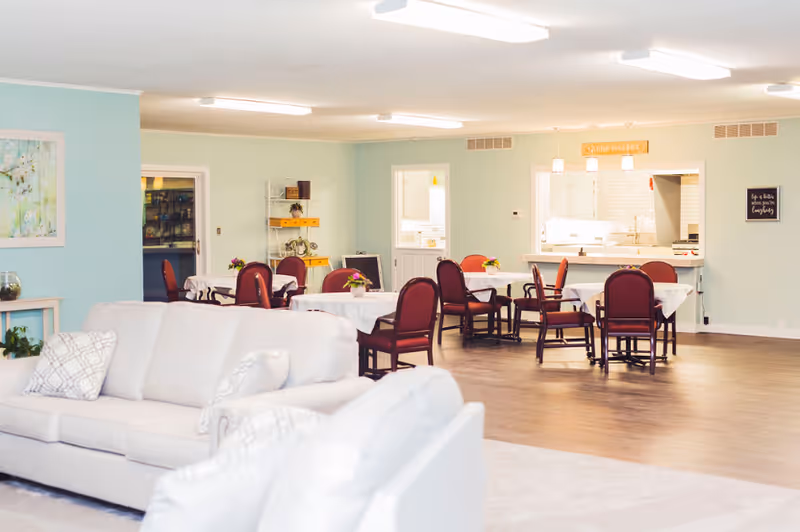 A bright and spacious common area with light blue walls, featuring several round tables with maroon chairs arranged around them. Each table has a small floral centerpiece. In the foreground, there are two light-colored sofas with patterned pillows. The background shows a kitchen pass-through window with a countertop and pendant lights hanging above it. The room has wooden flooring and soft overhead lighting.