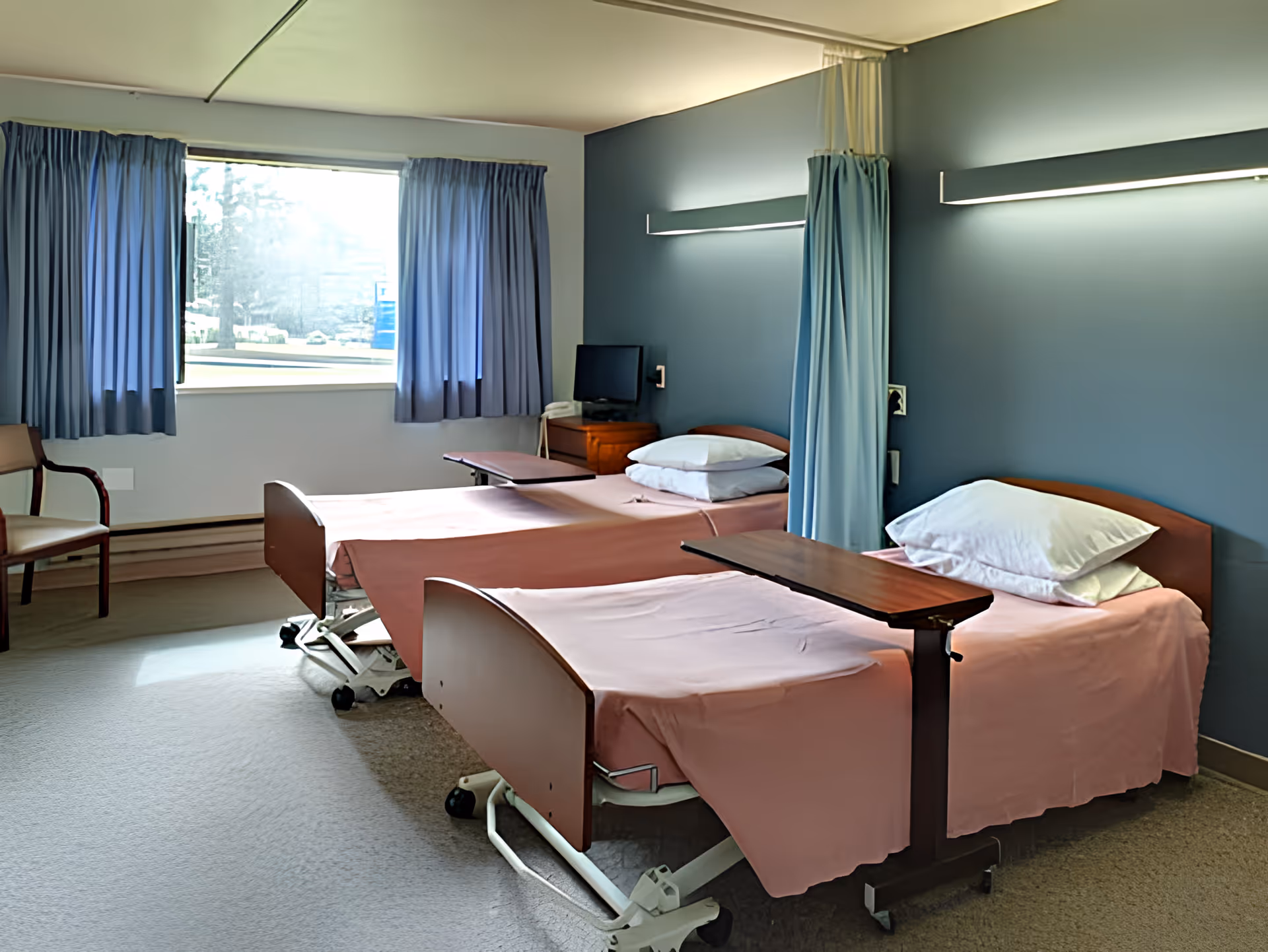 A healthcare facility room with two hospital beds covered in pink sheets, each with a pillow and an overbed table. The room has a large window with blue curtains, a wooden chair, a small TV on a wooden cabinet, and a blue privacy curtain between the beds. The walls are painted blue and white, and there is a long light fixture above the beds.