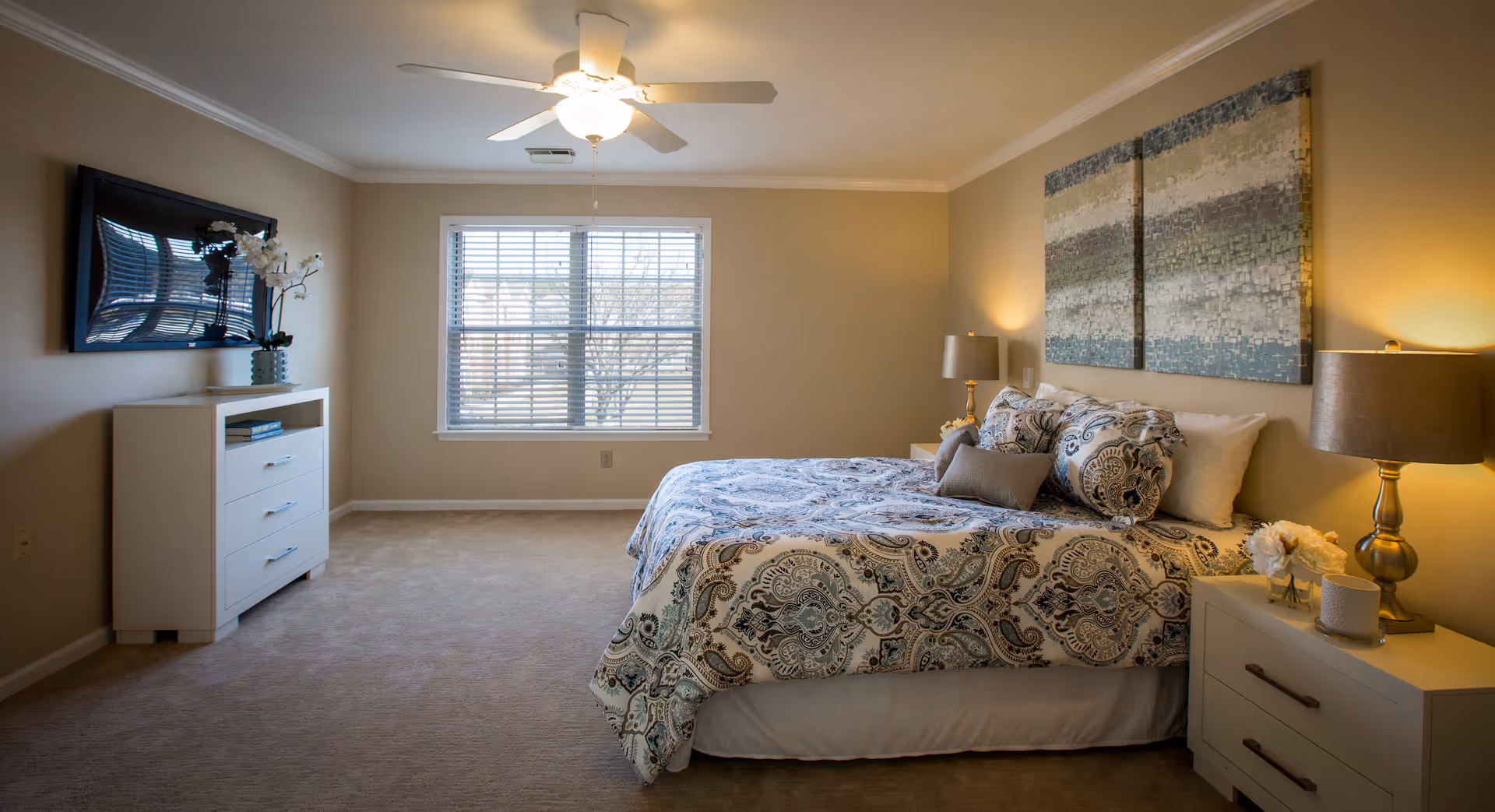 A well-lit bedroom featuring a large bed with patterned bedding and multiple pillows. On either side of the bed are nightstands with lamps and decorative items. A window with blinds is centered on the far wall, and a dresser with a vase of flowers and a mounted flat-screen TV is on the left side of the room. The walls are painted beige, and the carpet is light brown.
