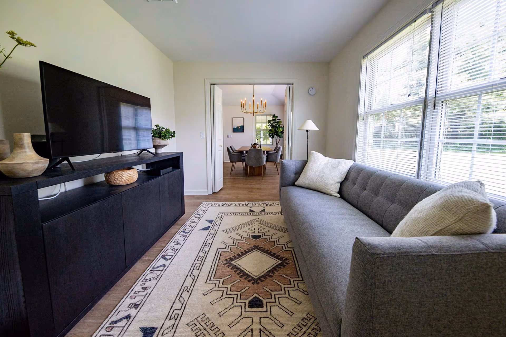 A bright living room with a large window on the right letting in natural light. There is a gray sofa with two light-colored pillows on the right side and a patterned area rug on the floor. On the left side, there is a black TV stand with a flat-screen TV and decorative items. In the background, through an open doorway, a dining room with a wooden table, gray chairs, a chandelier, and a large plant is visible.