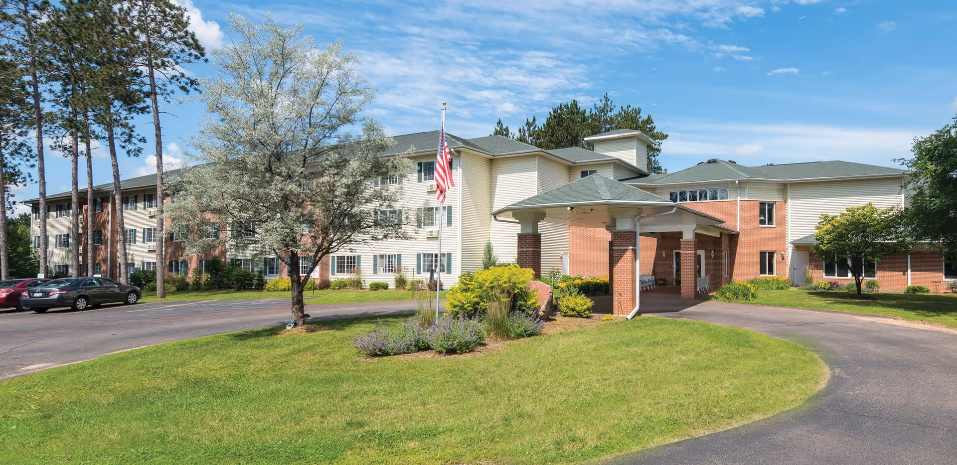 Front exterior of a multi-story senior living facility with a covered entrance, American flag, landscaped lawn, and parked cars.