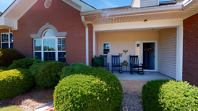 Front porch area of a residential building with two blue rocking chairs, a small table, and decorative items. The porch is surrounded by neatly trimmed green bushes and the building exterior features red brick and beige siding with a window on the left side.