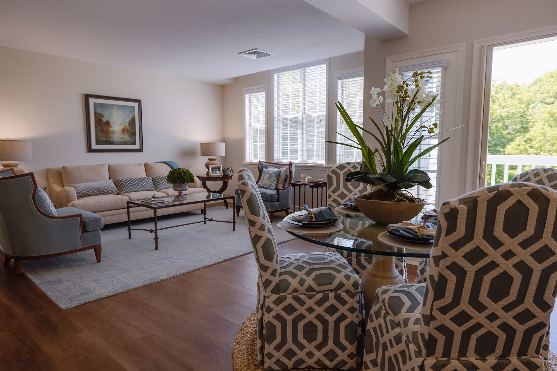 A bright and spacious living and dining area with large windows letting in natural light. The living room features a beige sofa with patterned cushions, two blue armchairs, a coffee table, and a framed painting on the wall. The dining area has a round glass table with four patterned upholstered chairs and a large floral centerpiece. Hardwood floors and neutral walls create a warm and inviting atmosphere.