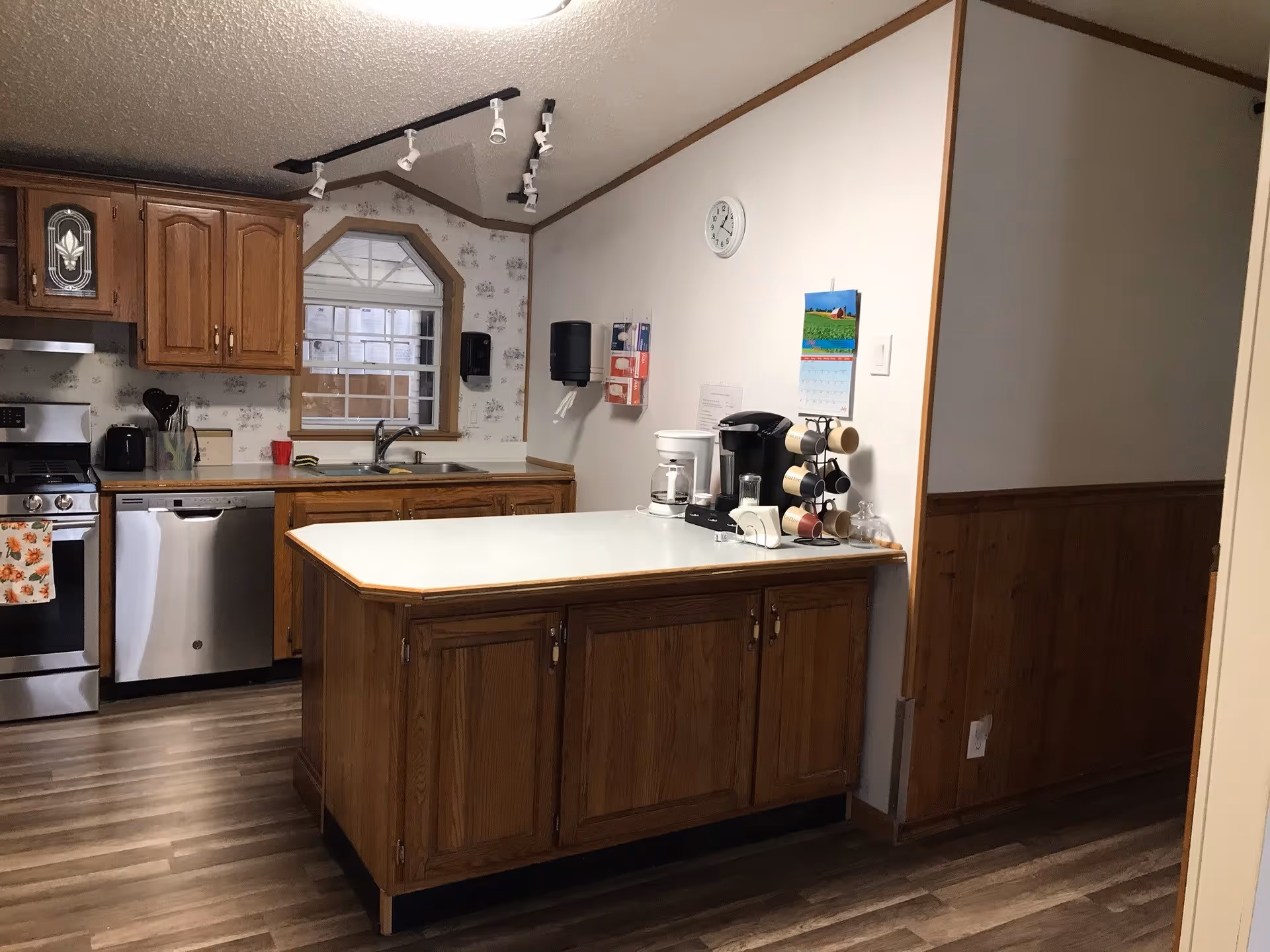 A kitchen area with wooden cabinets and a white countertop island. The kitchen features a stainless steel stove and dishwasher, a double sink under a window, and various kitchen appliances including a coffee maker and a Keurig machine on the island. The walls are partially covered with floral wallpaper and wood paneling, and there is a clock and a calendar on the wall.