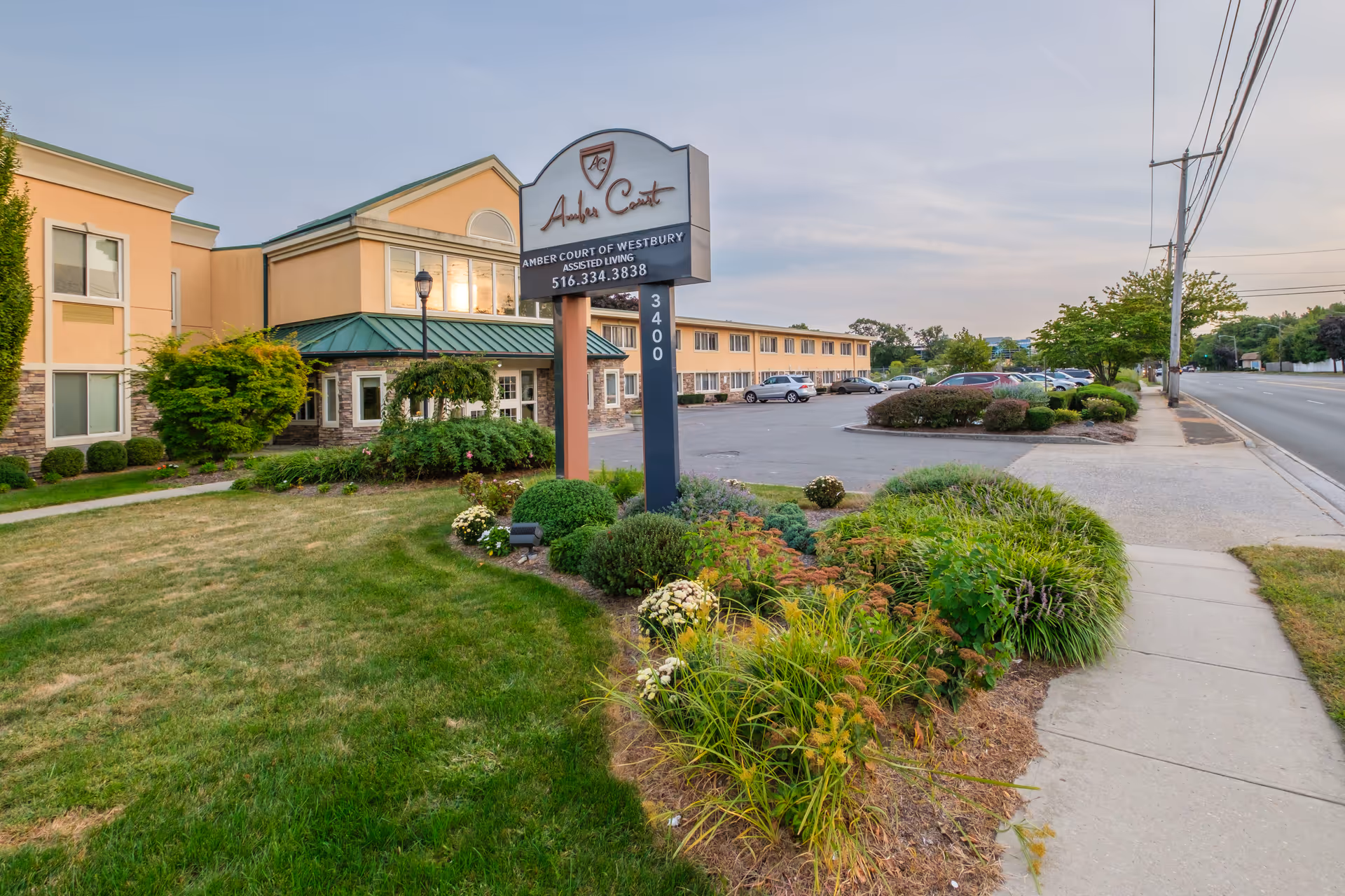 Exterior view of Amber Court of Westbury assisted living facility showing a two-story building with a green roof, landscaped garden with bushes and flowers, a parking lot with cars, a sidewalk, and a sign displaying the facility's name, phone number, and address.