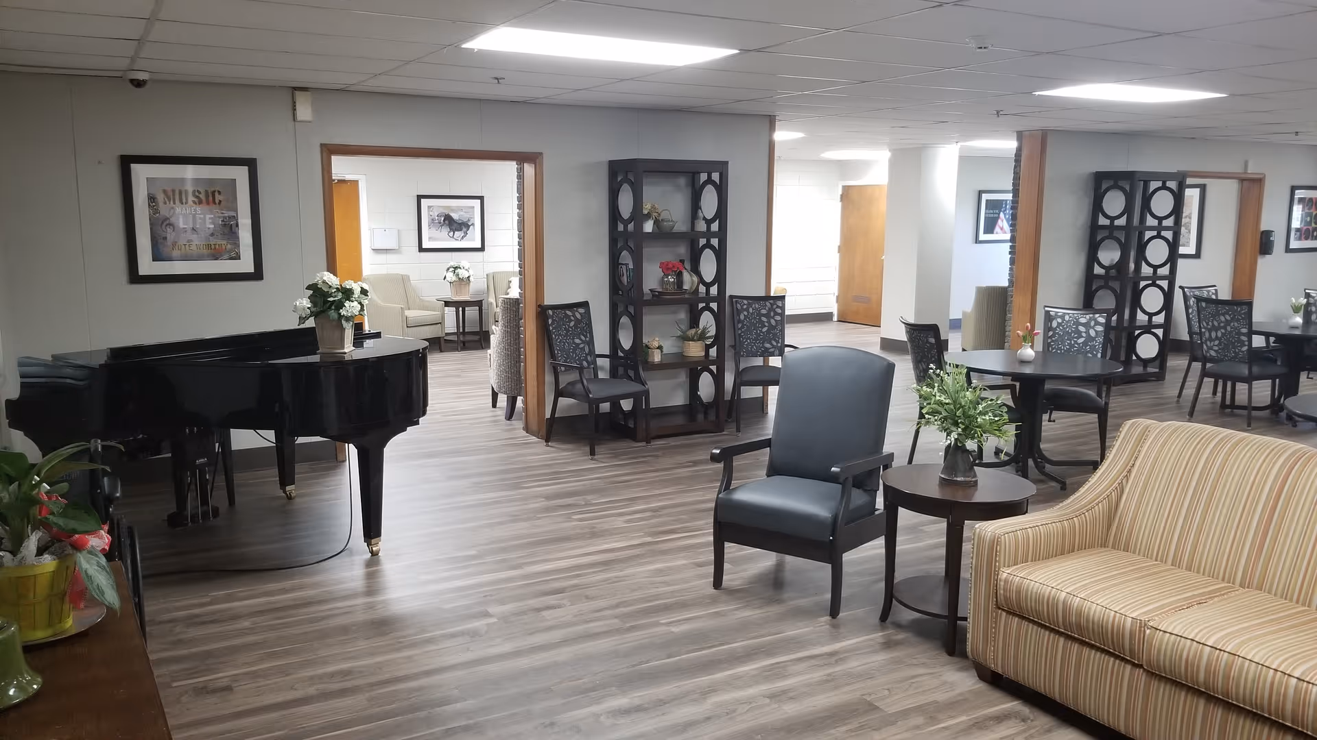 Spacious senior living common area with a grand piano, seating, tables, and decorative shelving.