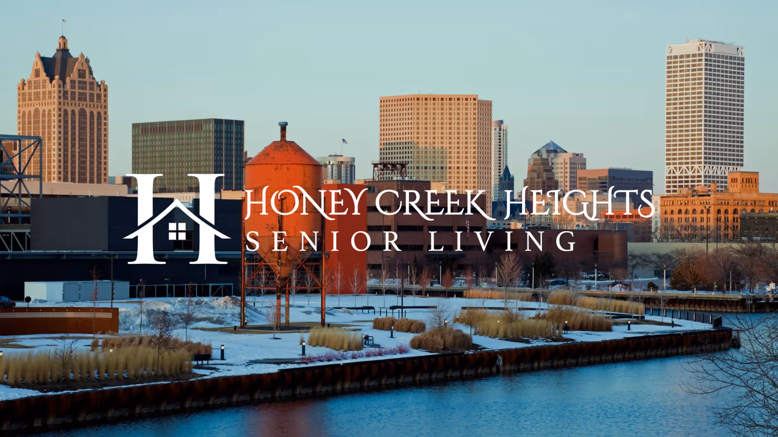 View of an urban waterfront area with a river in the foreground, landscaped banks with dry grasses, and a city skyline with various buildings in the background during daylight. The logo and text 'Honey Creek Heights Senior Living' are overlaid on the image.