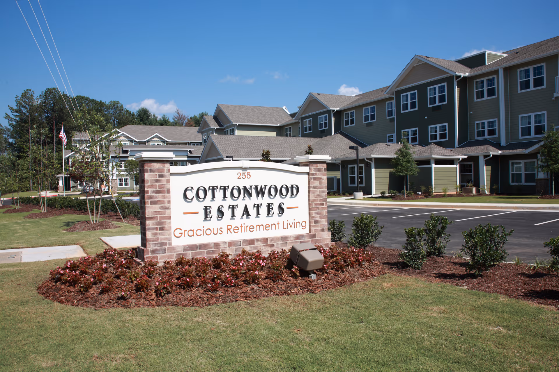 Exterior view of Cottonwood Estates Gracious Retirement Living facility with a large sign in front displaying the name. The building is multi-story with green siding and white trim, surrounded by landscaped grass, bushes, and a parking lot.