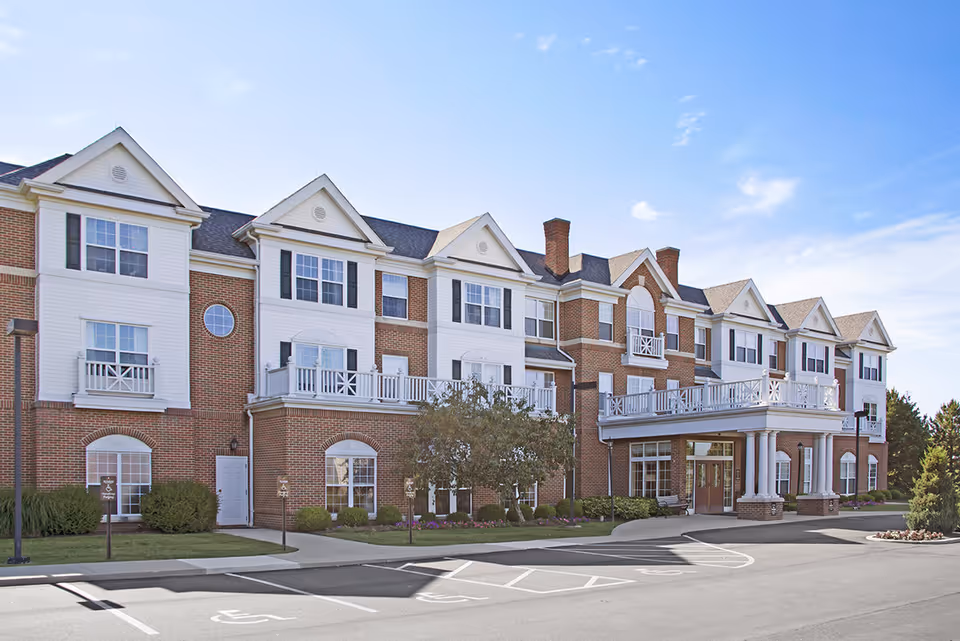 Exterior view of a large, multi-story senior living facility with brick and white siding, multiple windows, balconies, and a covered entrance with columns. The foreground shows a parking area with handicap parking spaces and landscaped greenery.