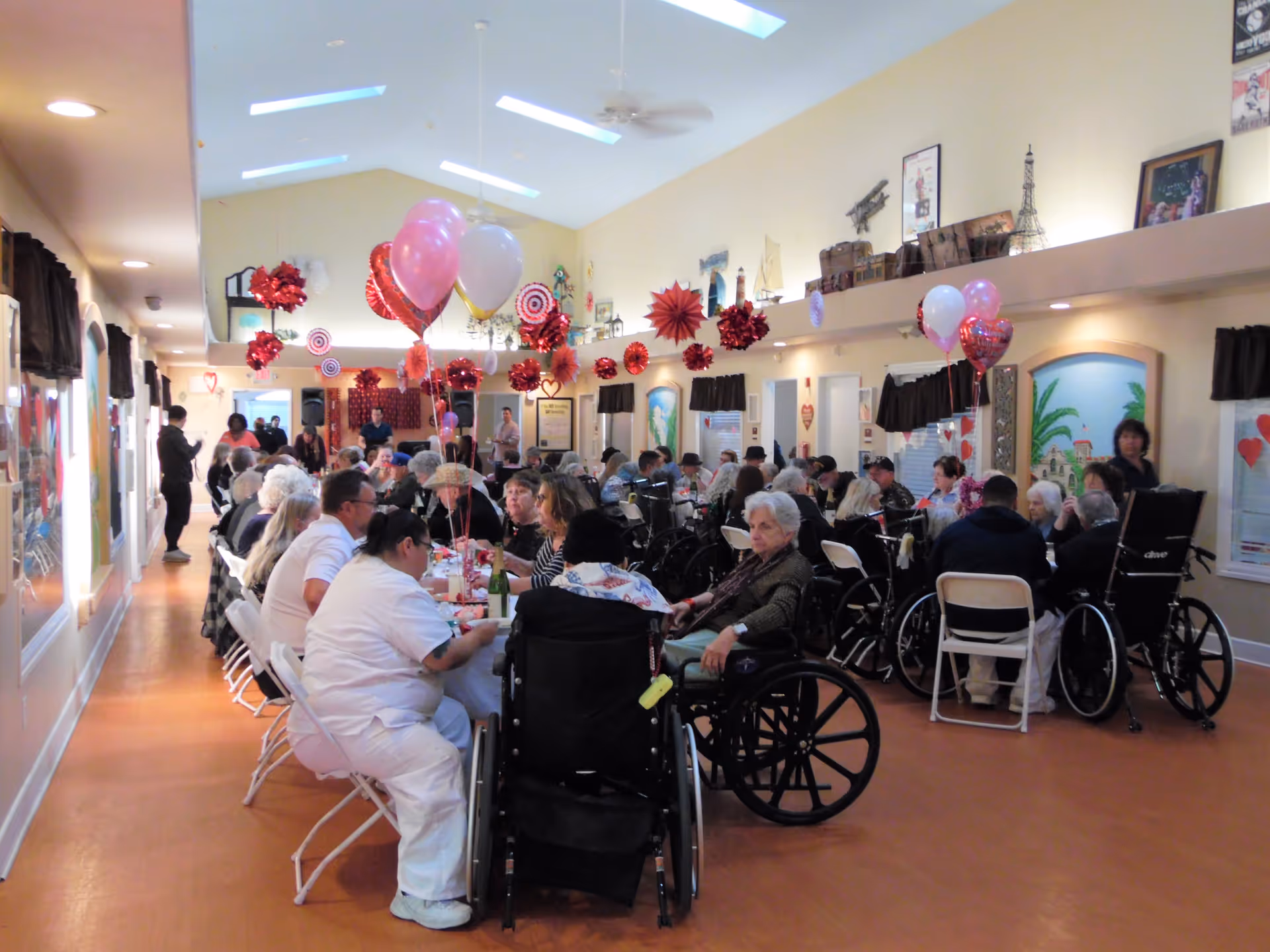 A large group of elderly people, some in wheelchairs, seated at long tables in a decorated common area. The room has high ceilings with skylights and ceiling fans. Red and pink heart-shaped balloons and paper decorations hang from the ceiling, suggesting a celebration or event. Staff members in white uniforms are attending to the residents.