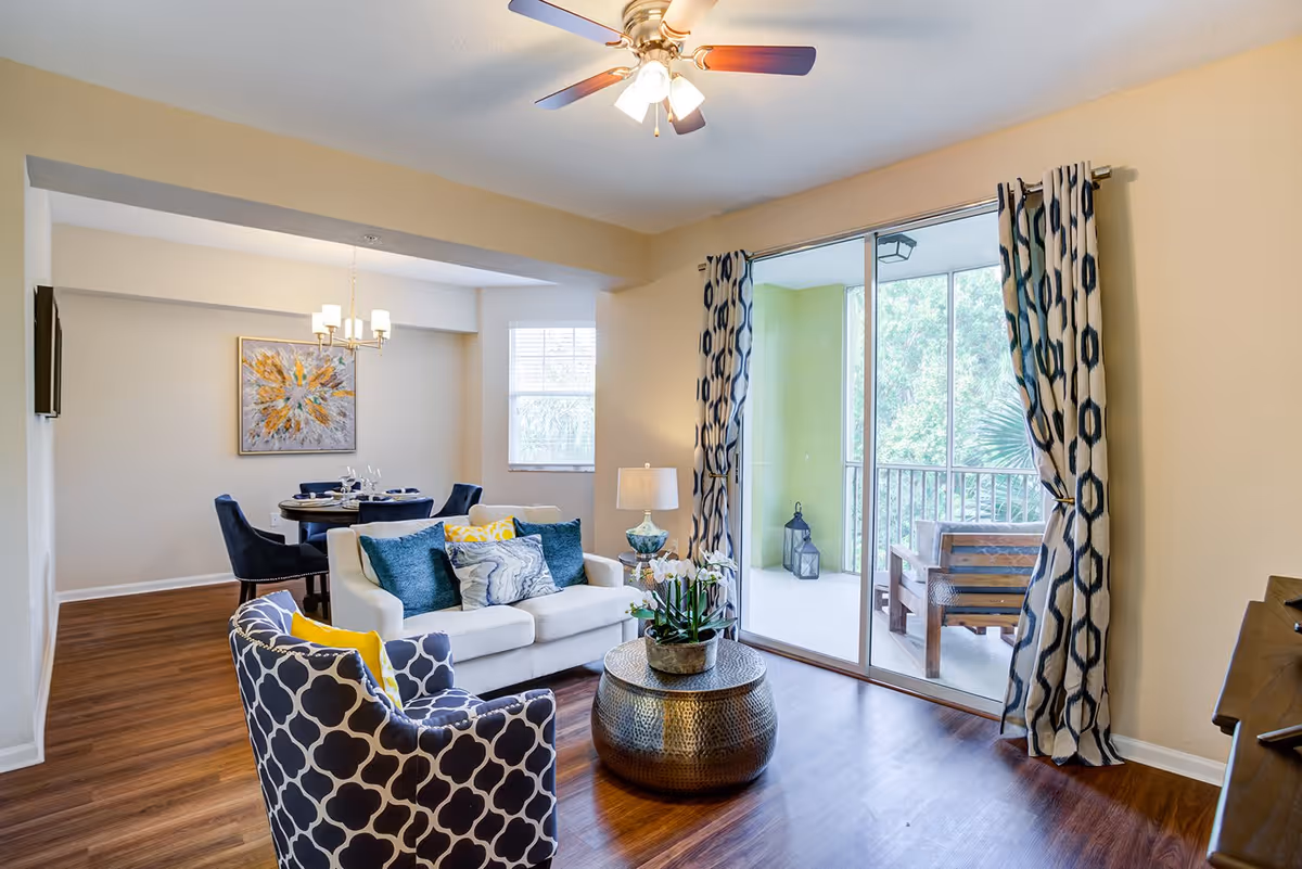 A bright living room with a white sofa adorned with blue and yellow pillows, a patterned armchair, and a round metallic coffee table with a potted plant. Behind the sofa is a dining area with a round table set for four and a modern chandelier. Large sliding glass doors with patterned curtains open to a balcony with wooden chairs and greenery outside.