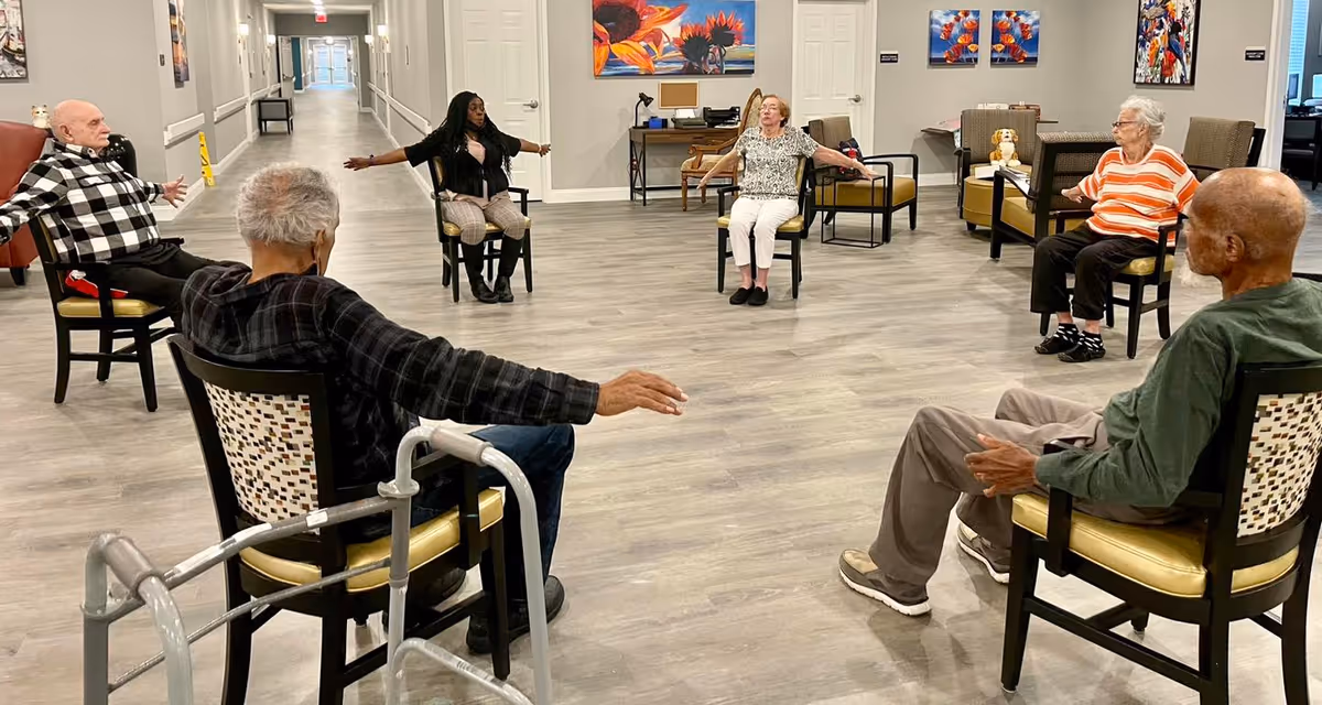 A group of elderly individuals seated in a circle on chairs in a spacious room, participating in a seated exercise or stretching activity led by a younger woman. The room has light-colored wood flooring, colorful artwork on the walls, and a long hallway visible in the background.