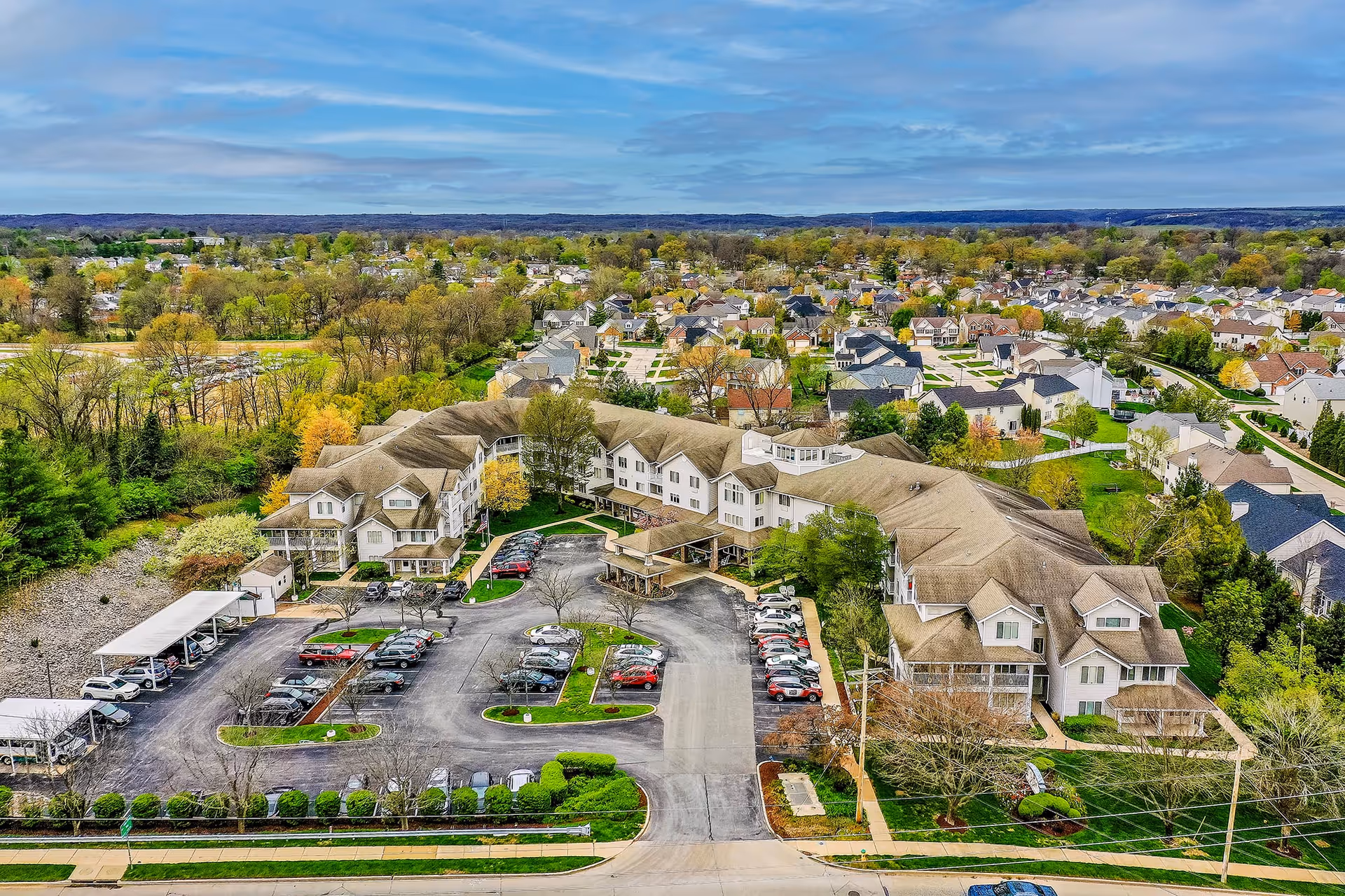 Aerial view of Solstice Senior Living at Fenton, showing a large senior living facility surrounded by parking lots, trees, and a residential neighborhood under a partly cloudy sky.