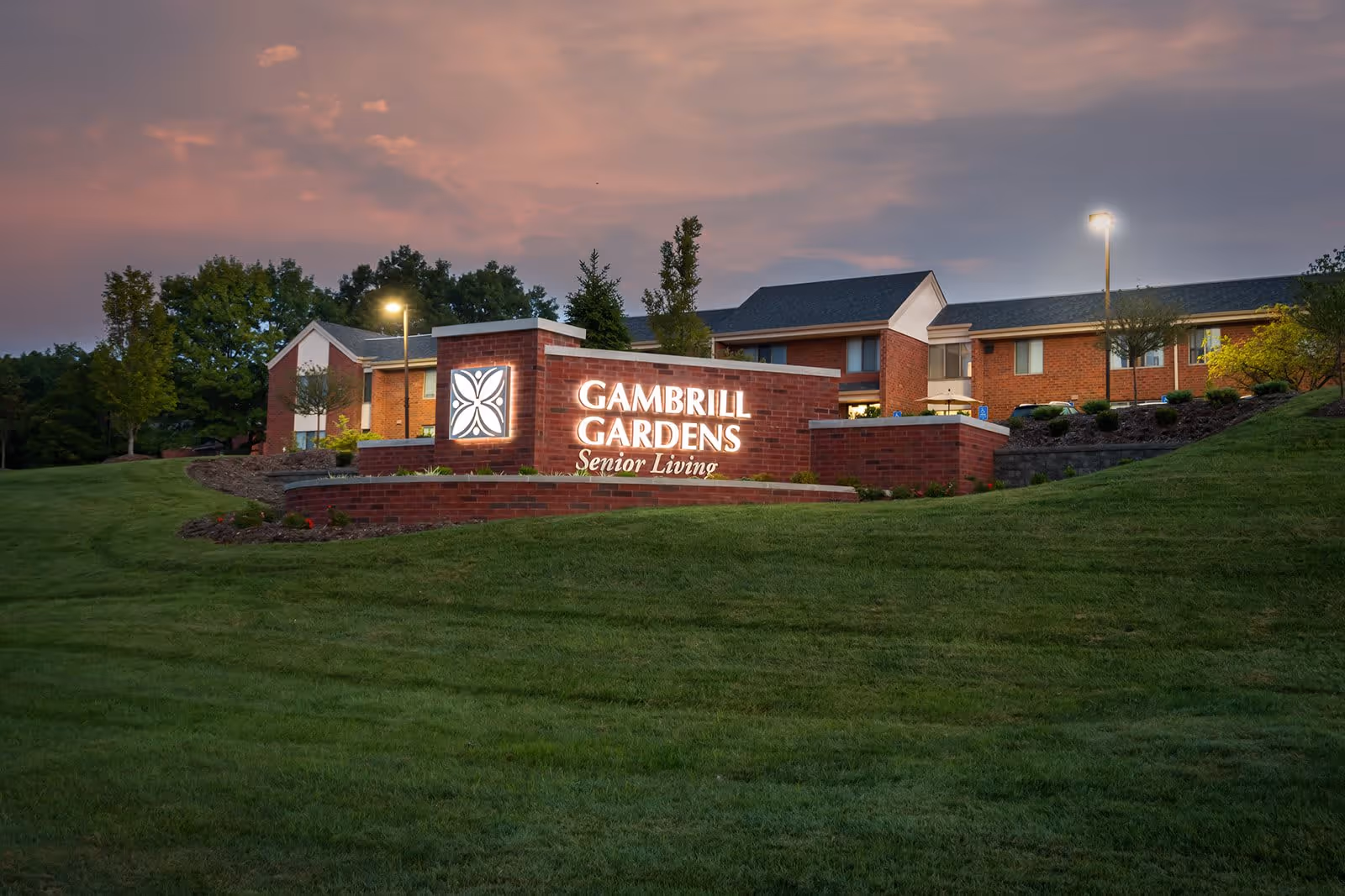 Exterior view of Gambrill Gardens Senior Living facility at dusk, showing a well-lit brick sign with the facility name and a background of a two-story brick building surrounded by green lawns and trees.