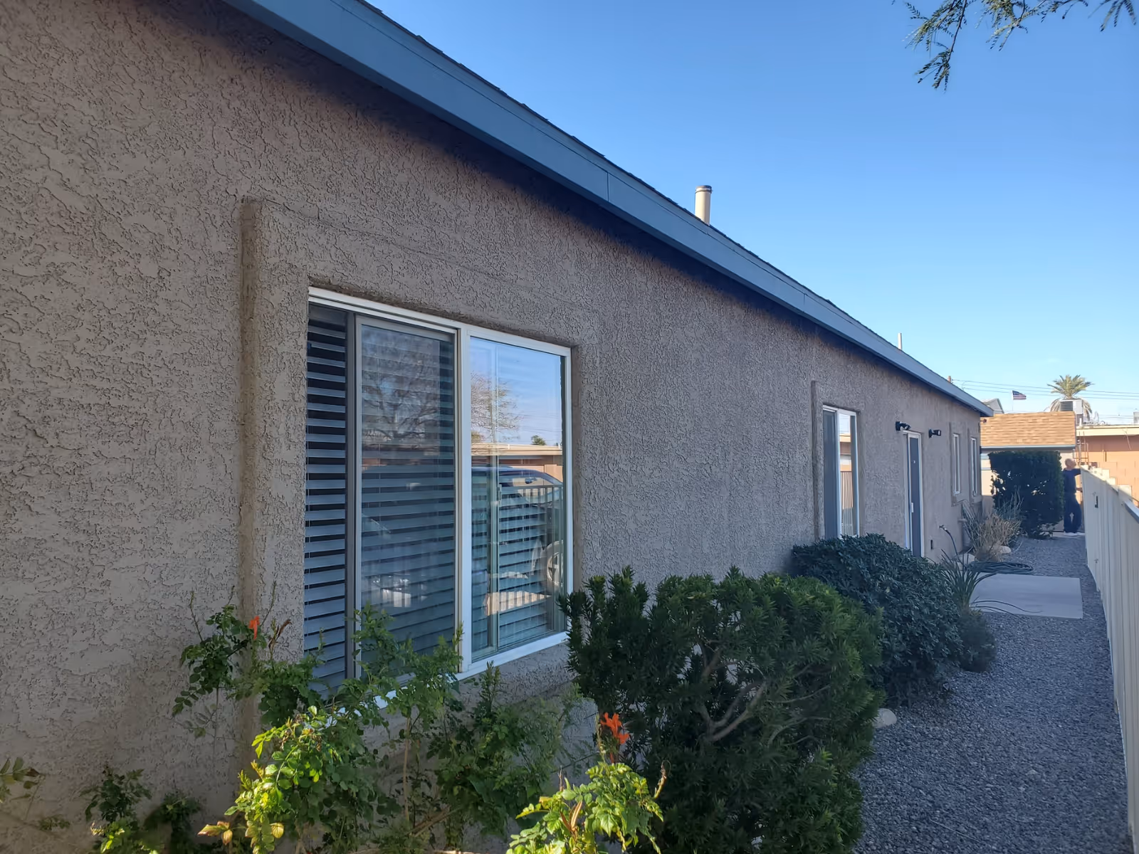 Side exterior view of a single-story stucco building with windows, shrubs, and a gravel walkway.