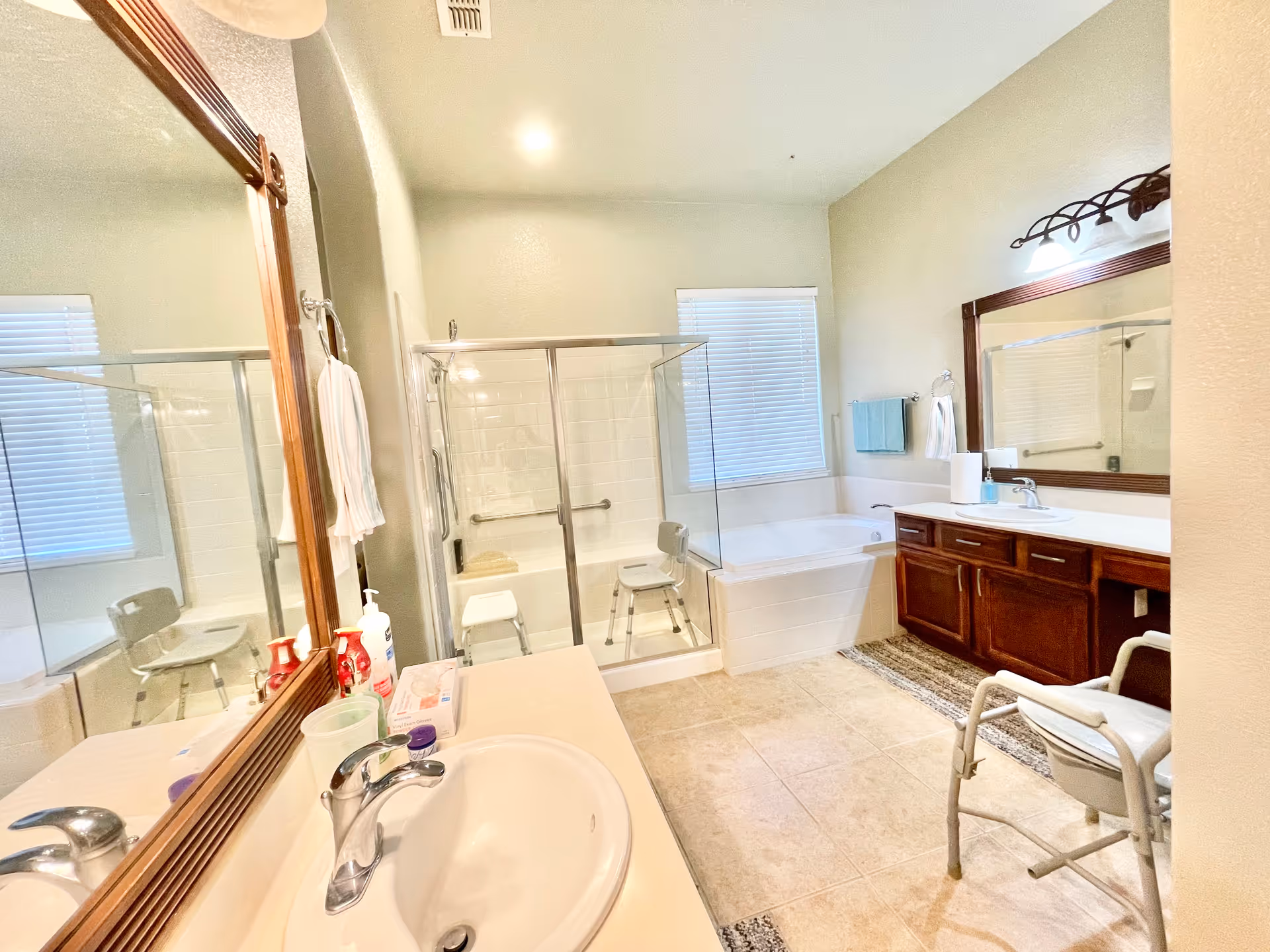 Bright and spacious bathroom with a large mirror above a sink, a glass-enclosed shower with a shower chair, a bathtub next to a window with blinds, wooden cabinetry with a countertop and sink, and a commode chair on the tiled floor.