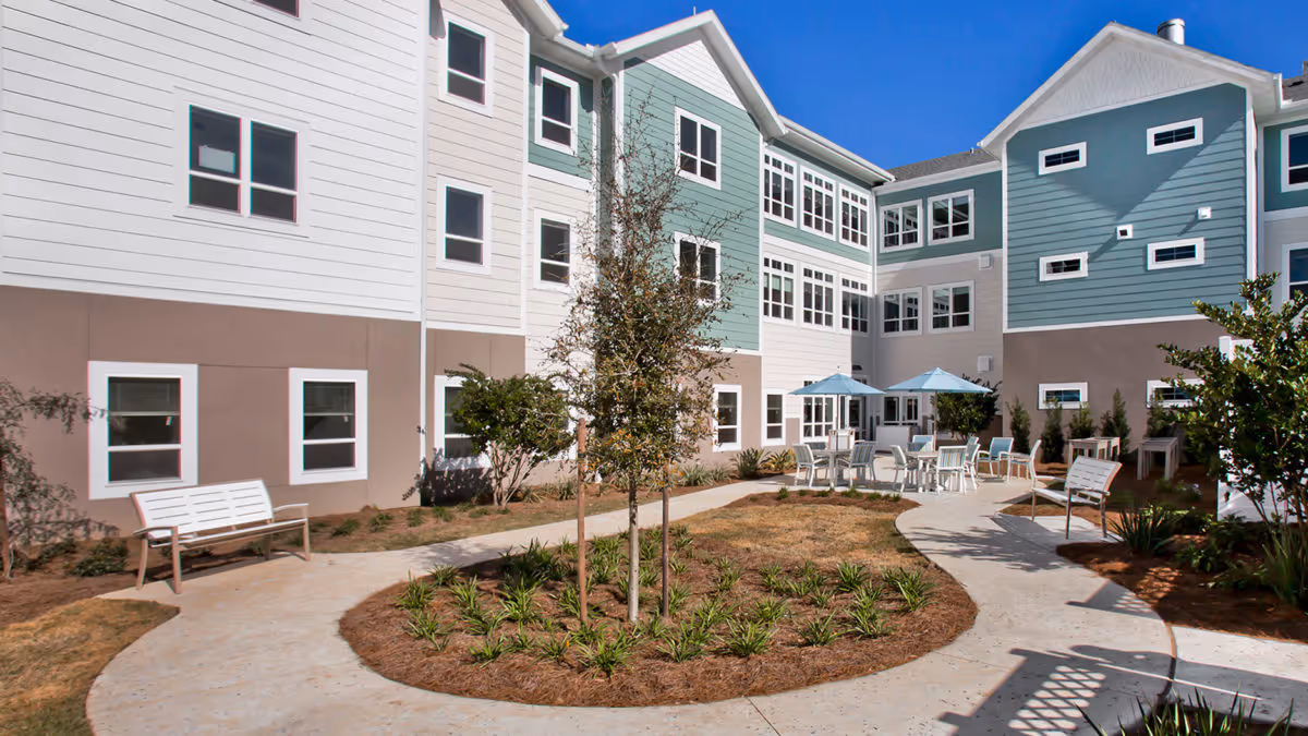 Sunny courtyard of a senior living building with winding walkways, benches, planted beds, and outdoor tables with umbrellas.