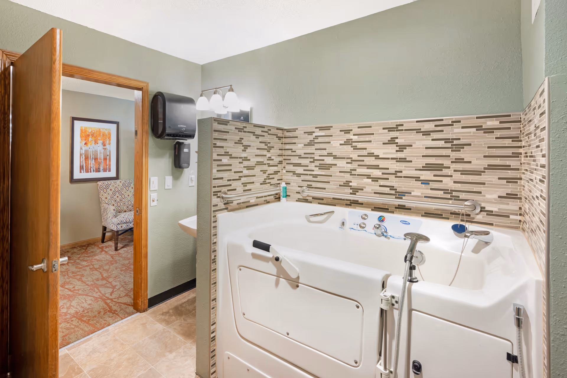 Walk-in accessible bathtub with mosaic tile backsplash in a bathroom, with an open door revealing a chair and framed artwork in an adjacent room.
