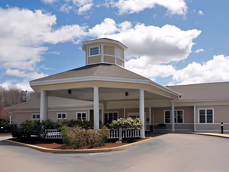Exterior front view of a single-story building with a covered entrance supported by white columns, surrounded by bushes and a clear blue sky with scattered clouds.