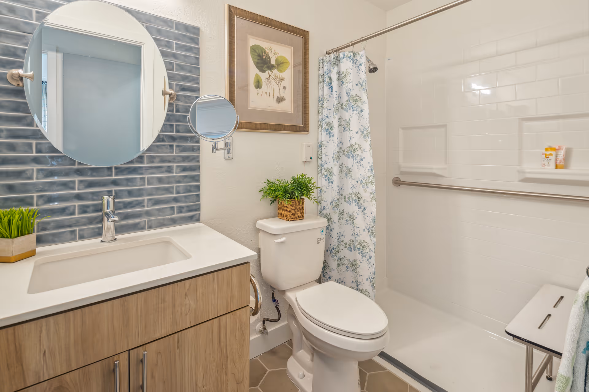 A bathroom with a white toilet, a wooden vanity with a white countertop and sink, a round mirror above the sink, and a blue tiled backsplash. There is a shower area with white tiles, a floral shower curtain, a grab bar, and a small bench. A framed botanical print hangs on the wall above the toilet, and a small potted plant is placed on the toilet tank.