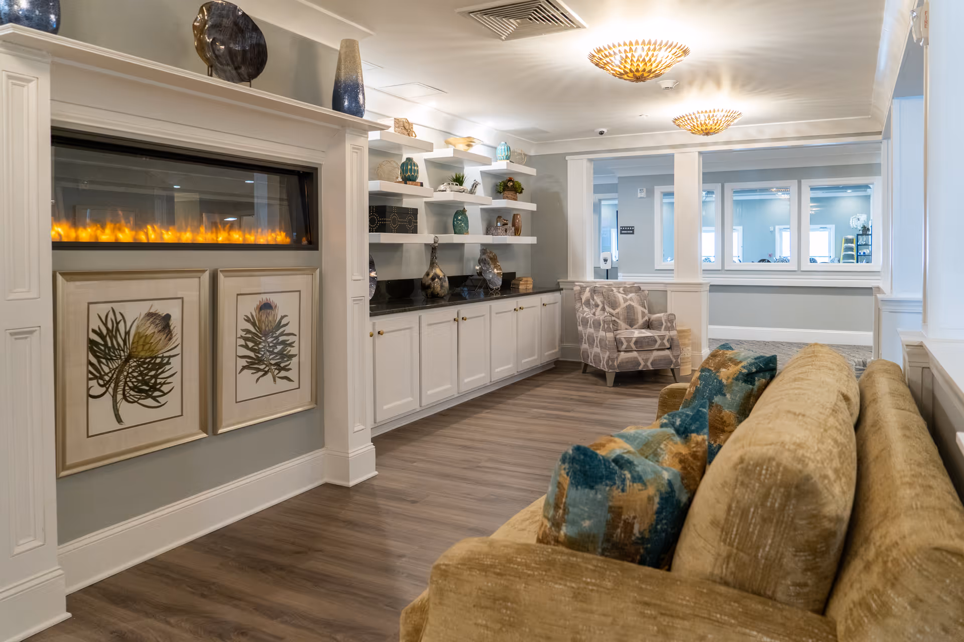 A cozy living room area in Arbor Ridge at Huntersville featuring a beige sofa with colorful cushions, a patterned armchair, a modern electric fireplace with two framed botanical prints below it, and white shelves displaying decorative vases and plants. The room has wood flooring and soft lighting from ceiling fixtures.