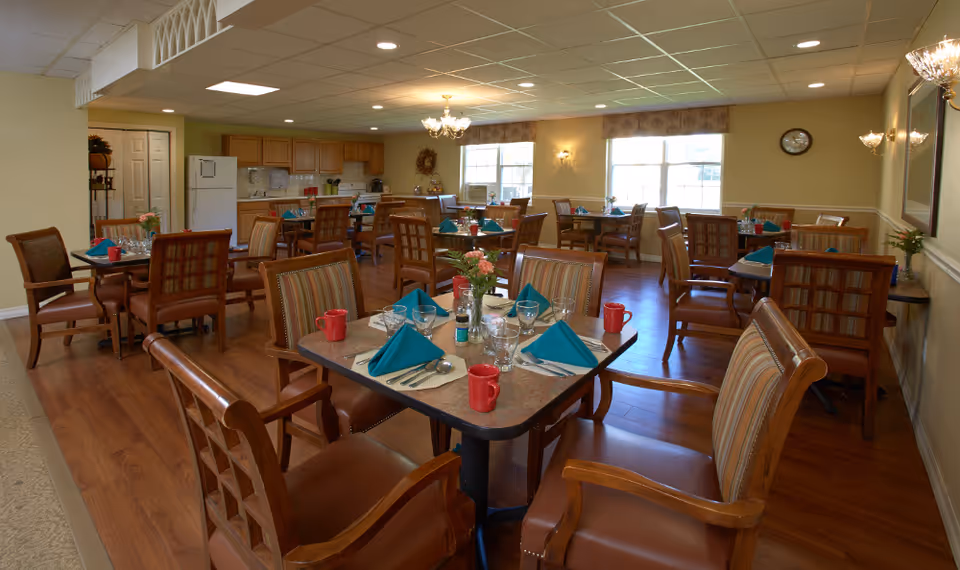 Dining room with multiple set tables and wooden chairs, place settings and centerpieces, and a small kitchen area in the background.